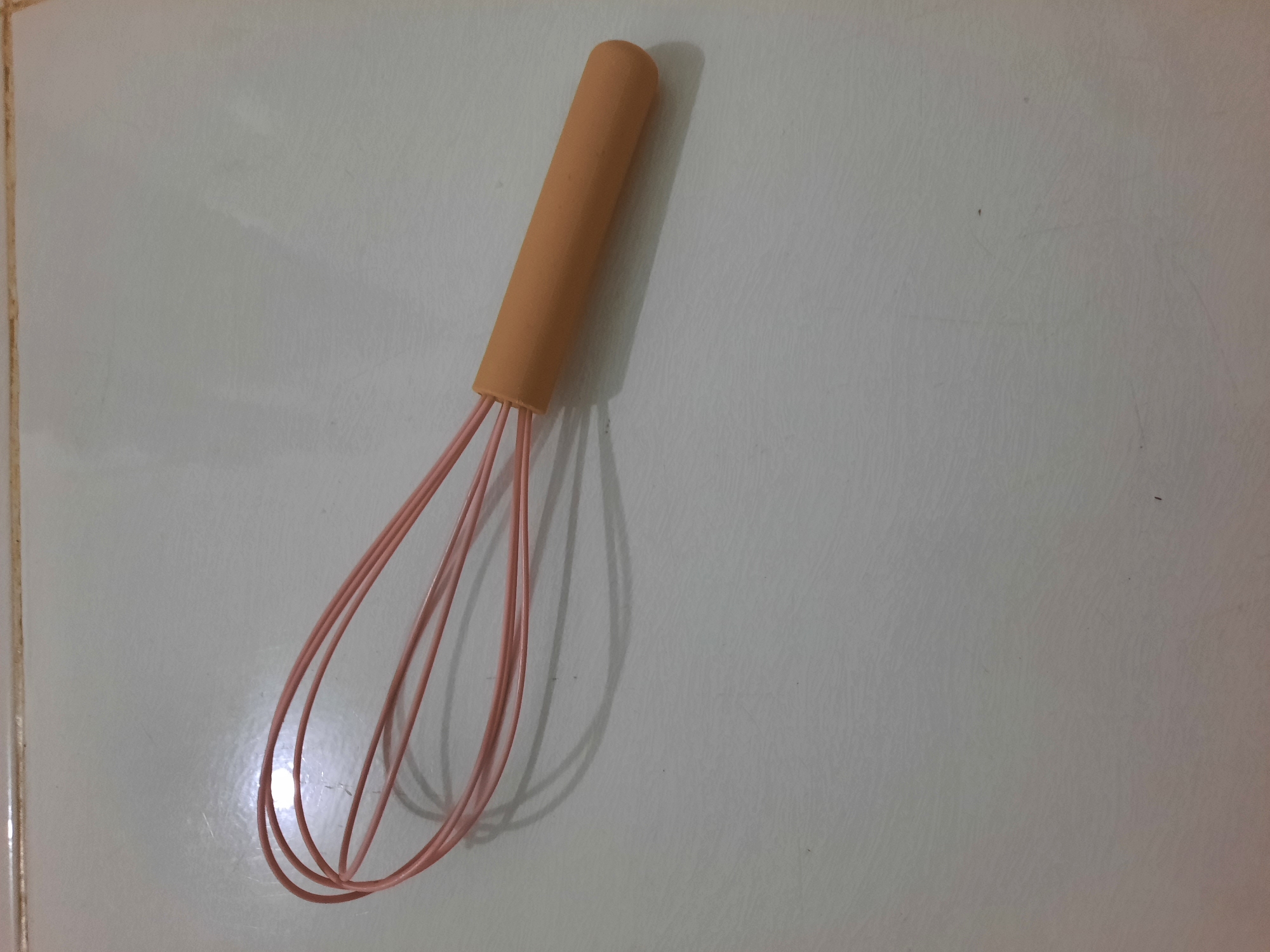 The image shows a pink whisk with an orange handle lying on a tiled surface. The tiles are light-colored with a subtle textured pattern. The whisk is positioned diagonally across the frame, with the handle on the right and the whisking loops on the left. The lighting suggests an indoor environment, casting a shadow from the whisk onto the tiles. The background is plain and unremarkable. There is no indication of the city of Lhokseumawe, Indonesia, or any specific environmental context that would suggest this location.