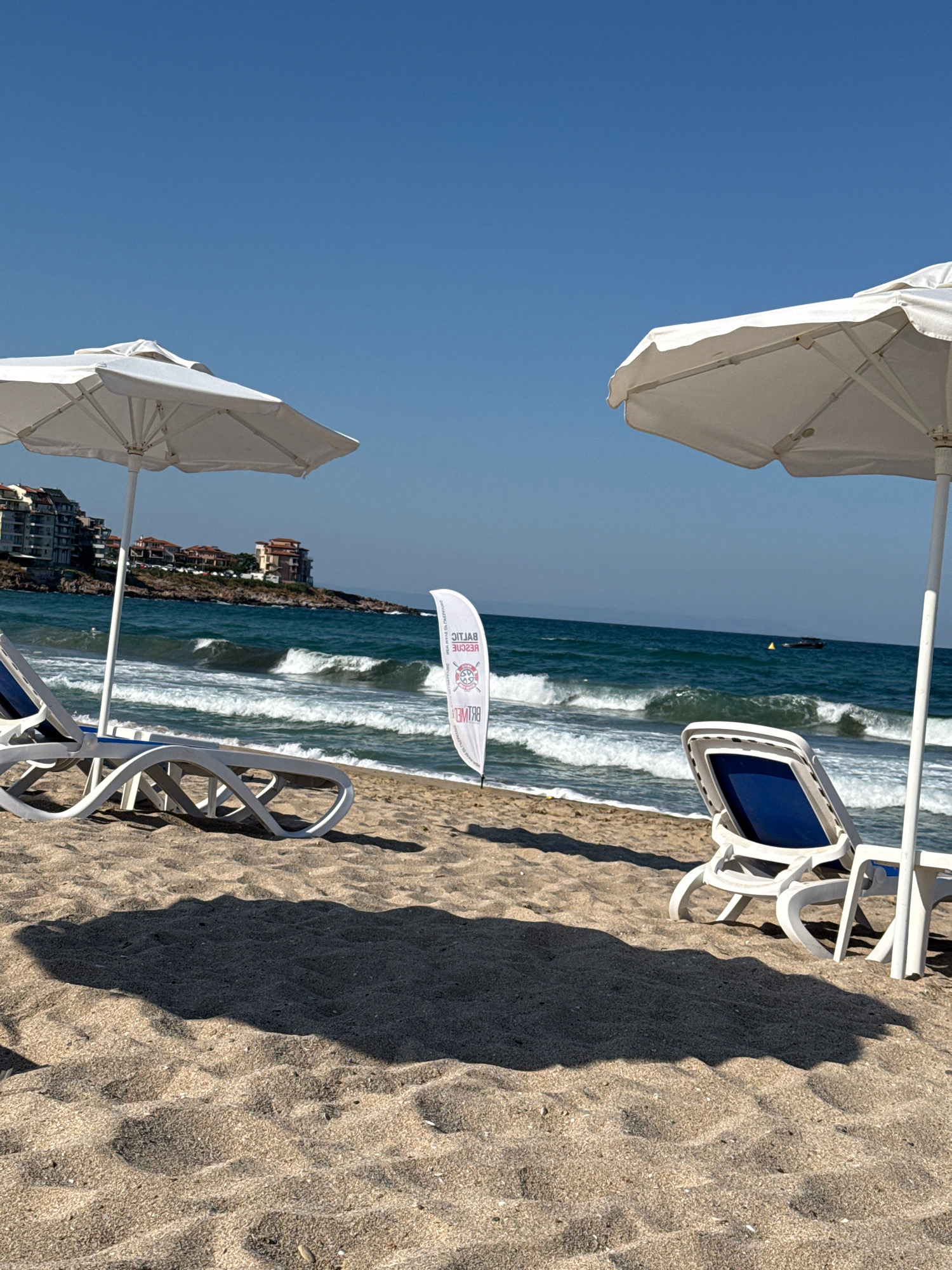 The image shows a view of a beach in Sozopol, Bulgaria, on a clear, sunny day. The foreground is dominated by sandy beach with shadows cast by two white umbrellas. Two empty white plastic beach chairs with blue cushions are visible, one on the left and one on the right, positioned under the umbrellas. In the mid-ground, the turquoise sea has moderate waves breaking on the shore. In the distance, along the coastline, there are several buildings, appearing to be apartment complexes or hotels, situated on a rocky outcrop. A white flag with red lettering and a circular logo stands upright on the sand near the center of the image. The sky is a clear, deep blue, and the horizon shows a faint line of distant land. A small boat is visible on the water in the far distance.