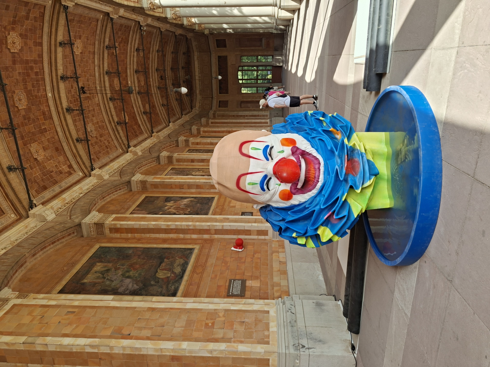 The image shows a large, colorful clown head sculpture displayed on a blue circular base filled with water. The sculpture is positioned in the foreground of a long, arched colonnade. The colonnade features ornate tiling on the ceiling and walls, with framed paintings adorning the upper walls. In the background, two individuals are visible, one standing and looking towards the end of the colonnade, and another walking away. The floor is paved with large tiles. The scene appears to be outdoors or in a covered, open-air space, with sunlight casting shadows on the ground. The location is identified as the Lichtentaler Allee, a public park in Baden-Baden, Germany, specifically in front of the Frieder Burda Museum.