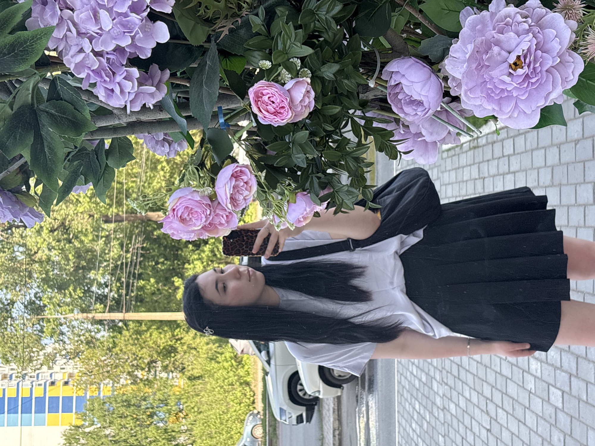 A young woman stands outside in a city, likely Izhevsk, Russia. She is dressed in a white t-shirt and a black skirt, with a black bag slung over her shoulder. She holds a cell phone in her hand, presumably taking a selfie. Her long, dark hair flows down her back. Above and around her are artificial flowers in shades of pink and purple, creating a floral archway. The background includes trees, a building with blue and yellow accents, and a white car. The ground is paved with light grey bricks.