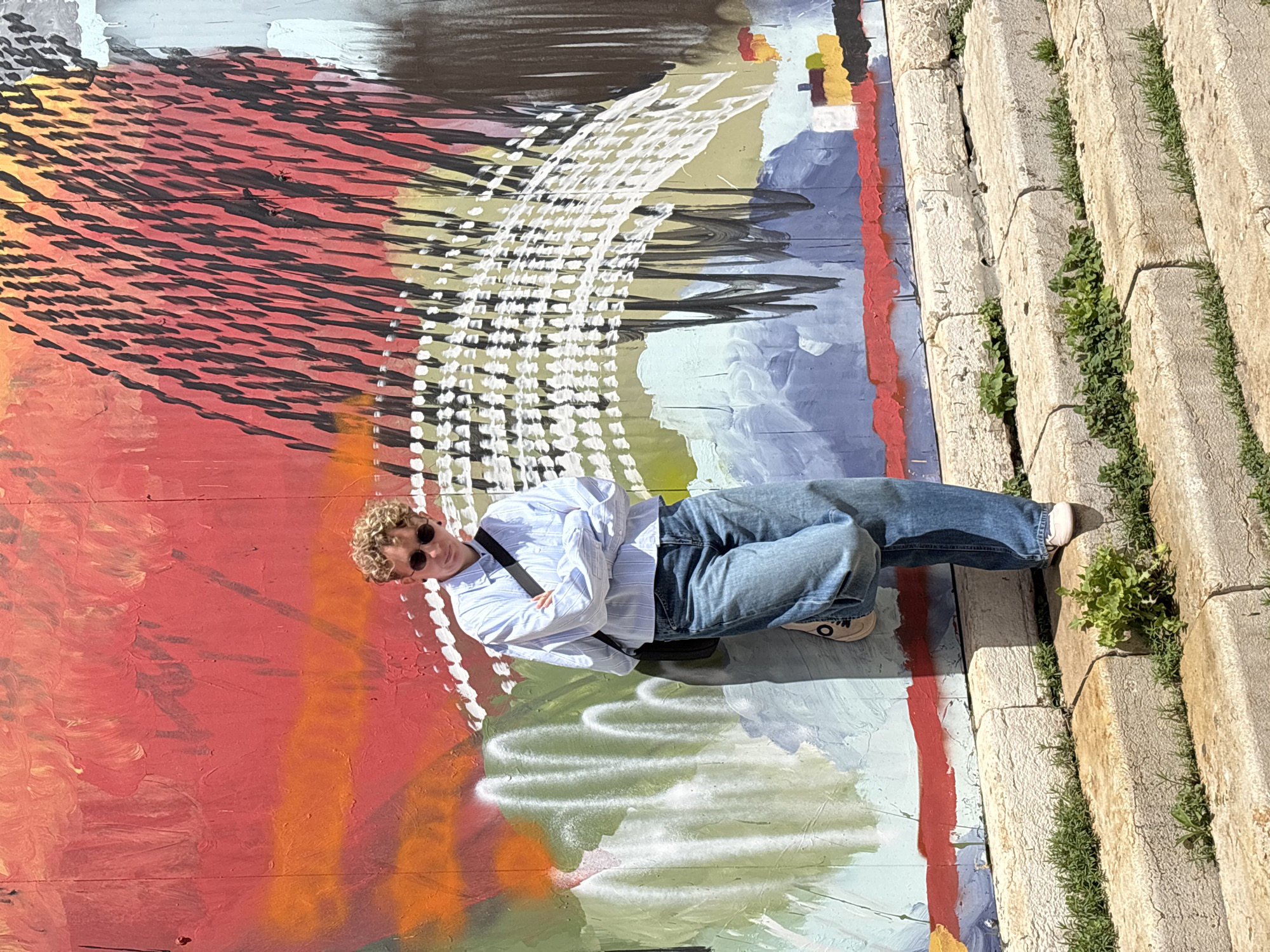 A person is lying on a colorful painted wall next to stone steps with plants growing in the cracks. The person has fair skin and curly, blonde hair and is wearing sunglasses, a long-sleeved blue-and-white-striped button-up shirt, blue jeans, and white shoes with black markings. They have a black strap across their chest. The wall is painted with red, orange, white, green, grey, blue and black. The stone steps are beige, and the plants are green. The photo appears to have been taken outdoors in natural sunlight. This photograph was taken in the city of Tel-Aviv, Israel.