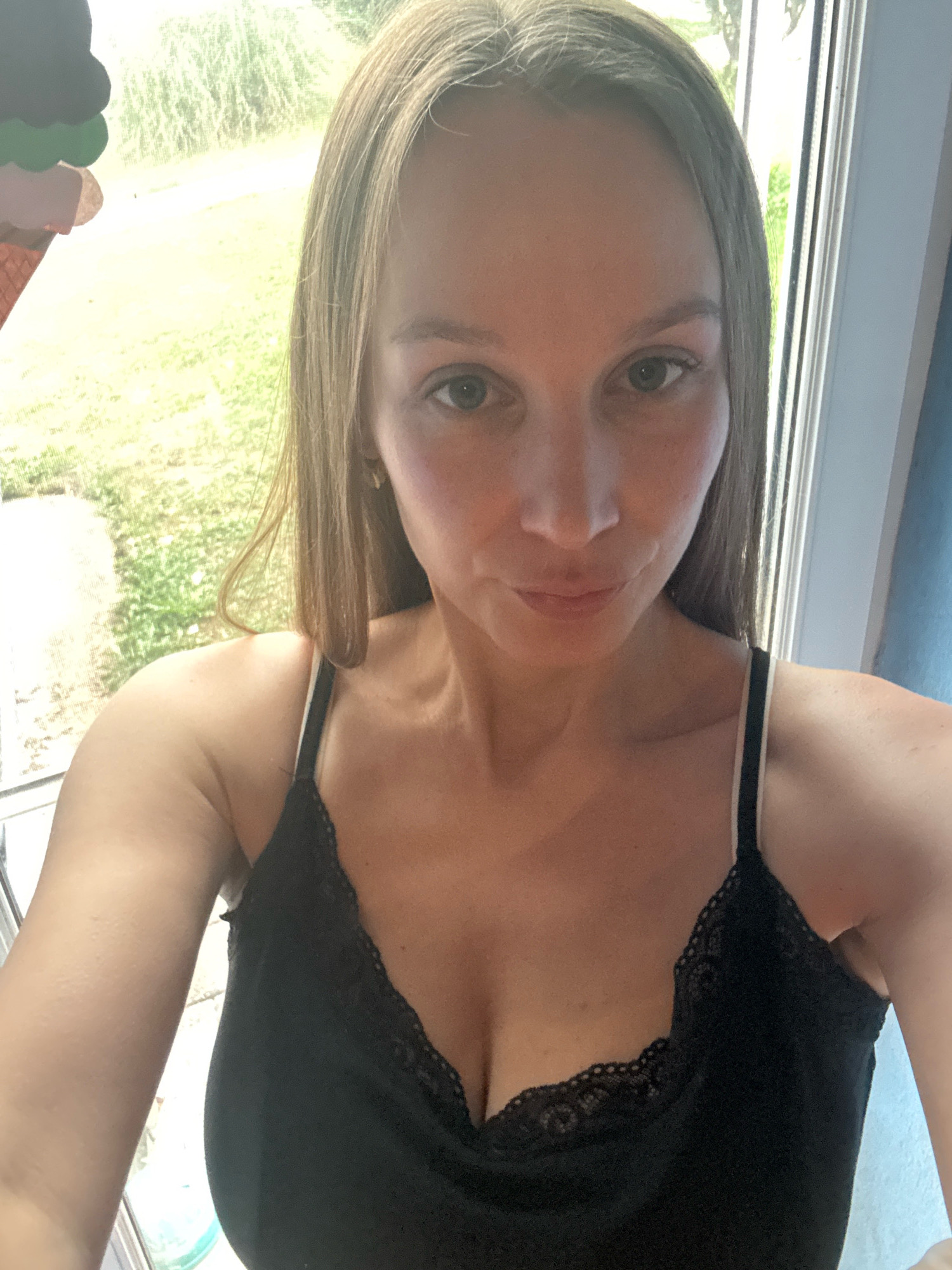 This is a selfie of a fair-skinned woman with blonde hair, taken indoors, likely in Überherrn, Germany. She is wearing a black lace-trimmed camisole. The background shows a window with a view of greenery outside. To the left of the frame, a portion of what appears to be a colorful decoration, possibly representing an ice cream cone, is visible. The lighting suggests it is daytime.