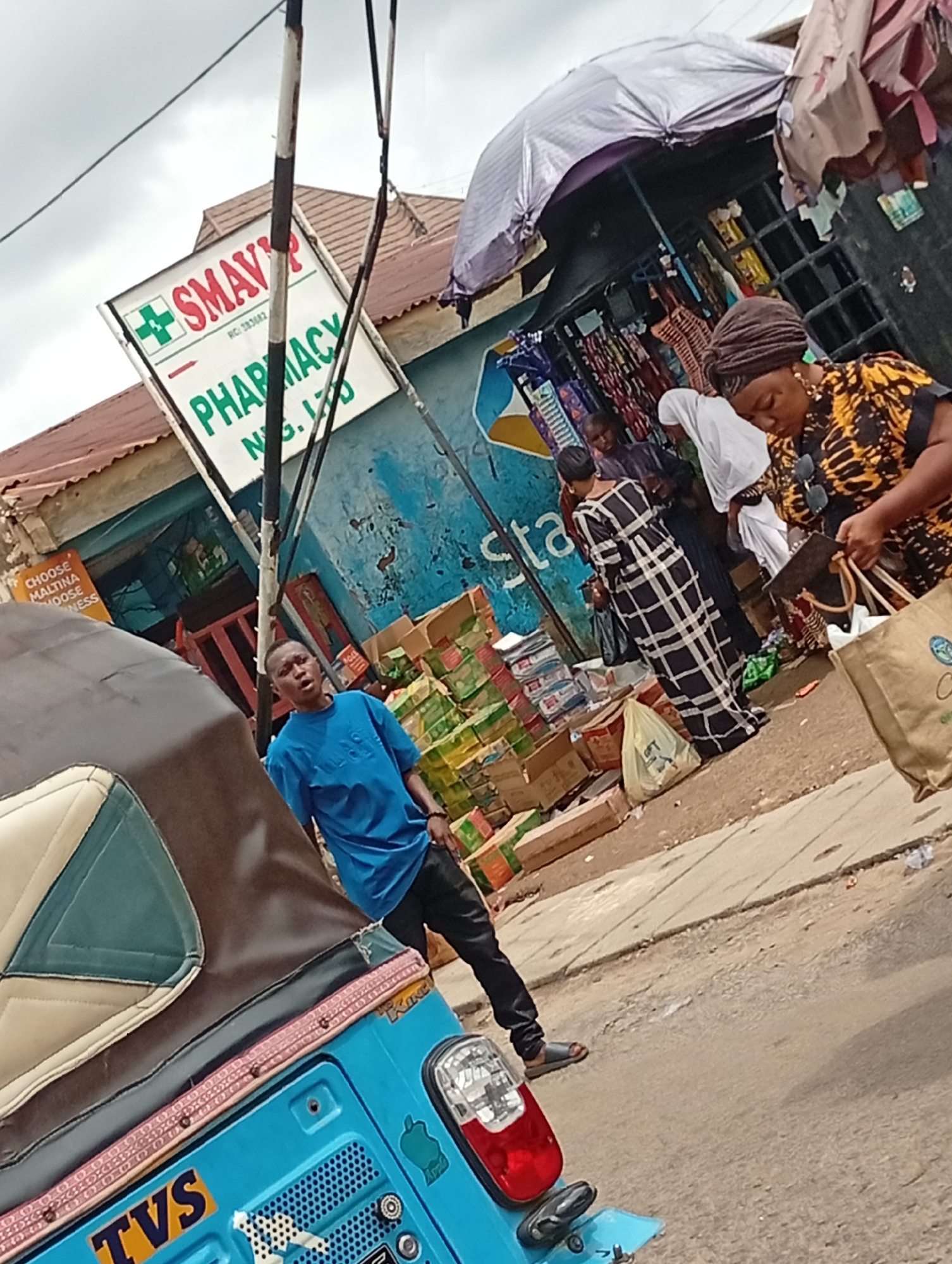 The image depicts a street scene in Lokoja, Nigeria, with a blue TVS tricycle taxi prominent in the foreground. In the background, several people are interacting near a shop, including women in patterned dresses. To the left is a sign for "SMAY'P PHARMACY." The area appears to be a bustling commercial street with boxes of goods displayed outside the shop.