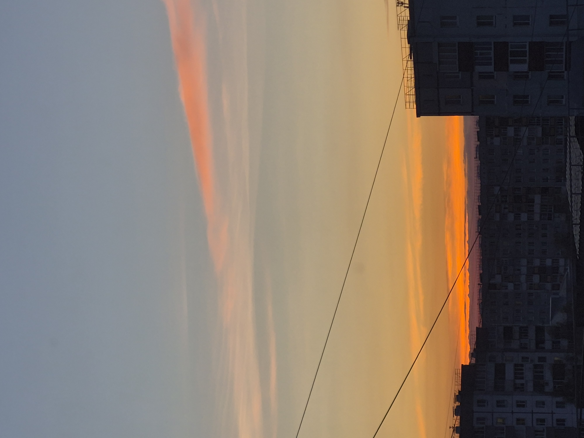 The image captures a sunset scene in Tobolsk, Russia. The sky transitions from a pale blue on the left to a vibrant orange near the horizon. Light orange clouds streak vertically across the sky. Silhouetted apartment buildings line the right side of the image, their windows visible. Electrical wires stretch across the view. The photograph is taken from a low angle, possibly street level, looking upwards.