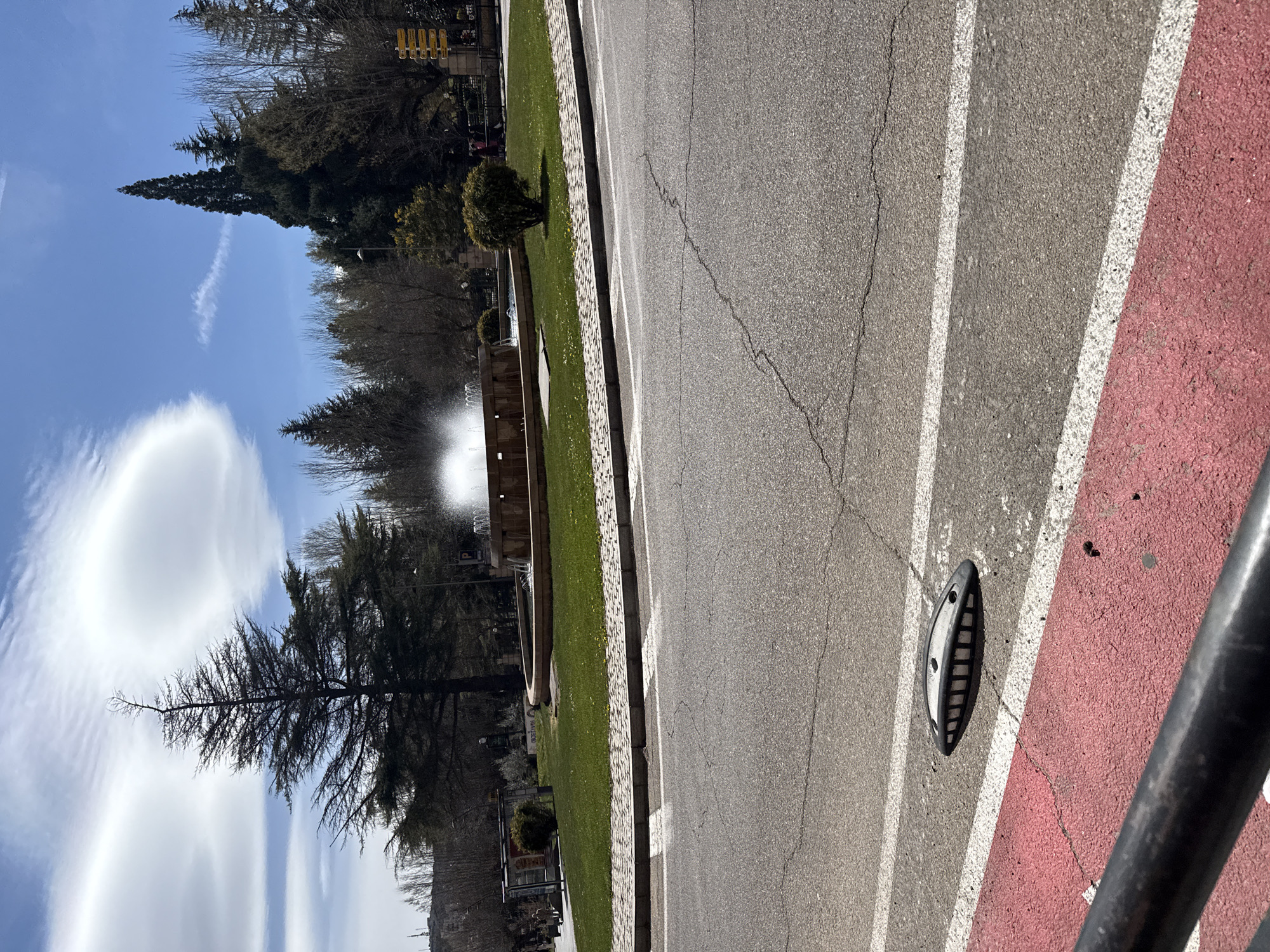 The image captures a street scene in Soria, Spain, featuring a road with a red-colored lane running alongside it. The road is marked with white lines. A raised circular drain cover is visible on the road. Beyond the road, there is a grassy area bordered by trees and a fountain. The sky is blue with scattered clouds. The photograph seems to be taken from an elevated position, potentially near a railing.