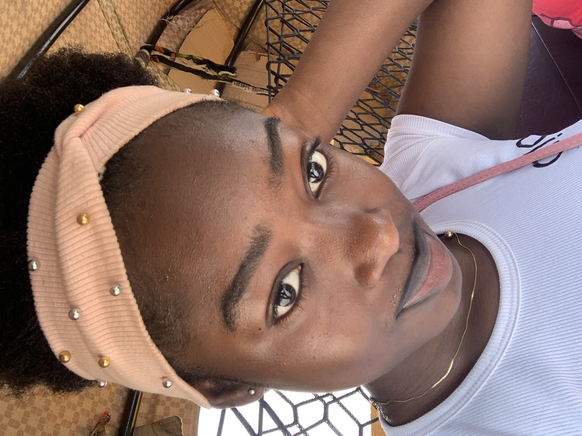 A young Black woman is seated outdoors, looking at the camera with a slightly tilted head. She wears a light pink headband adorned with gold and silver studs, framing her dark, curly hair. She has on a white ribbed tank top and a delicate gold chain necklace. A thin pink purse strap is visible. She is sitting on a black wire mesh chair, and the background includes a patterned cushion and what appears to be an outdoor setting. The location cannot be determined with certainty to be Rafin Par, Nigeria.