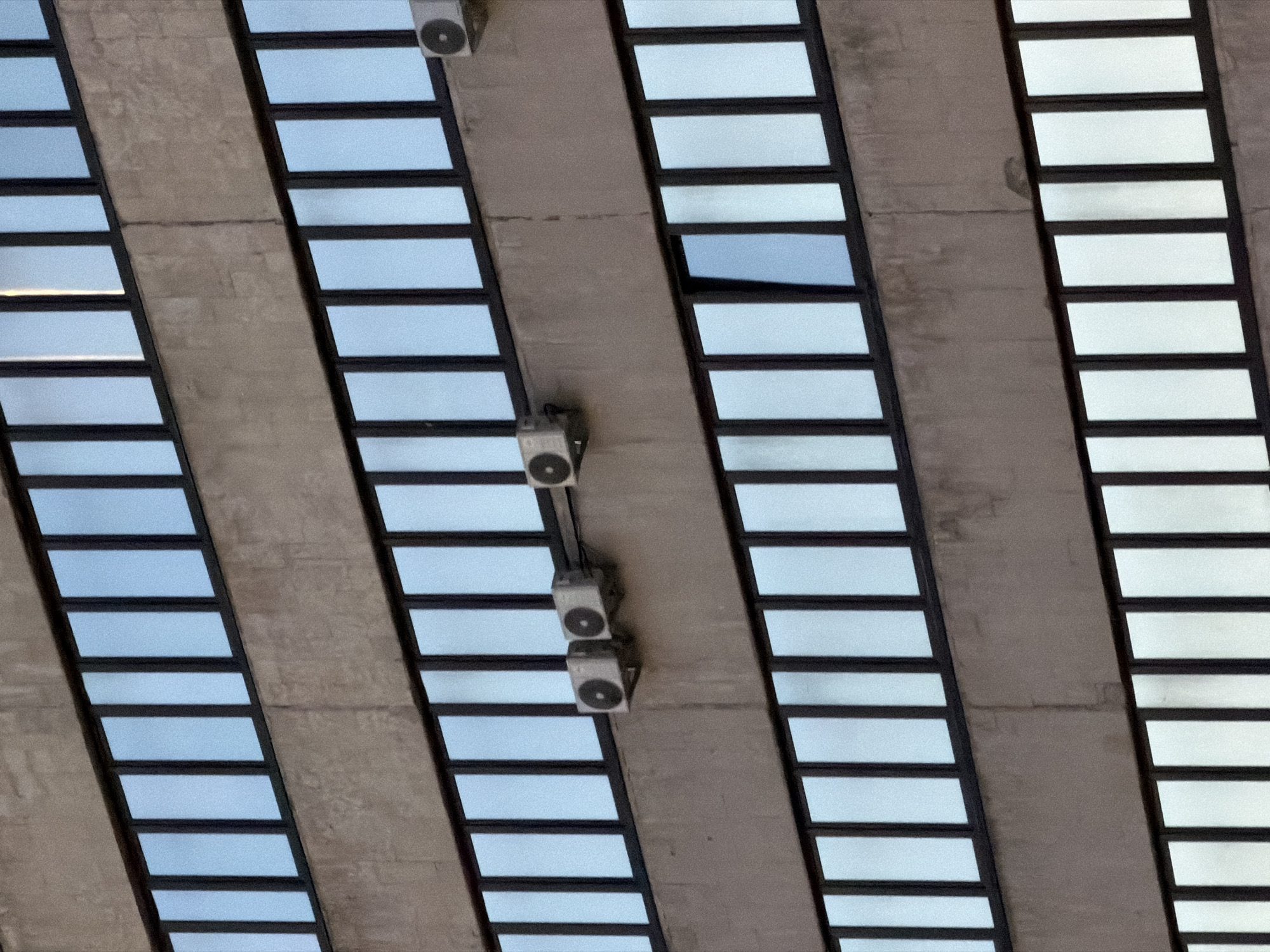 The image shows a close-up, eye-level view of the exterior of a Brutalist-style building, likely an office or apartment complex, with rows of reflective windows and a concrete facade. Several outdoor air conditioning units are mounted on the facade between the window levels. The windows are a light blue color, reflecting the sky, and are divided by dark vertical frames. The concrete appears weathered and textured. The building is located in Moscow, Russia, and there are no people visible in the image. The time of day is unclear, but the lighting suggests it is daytime.