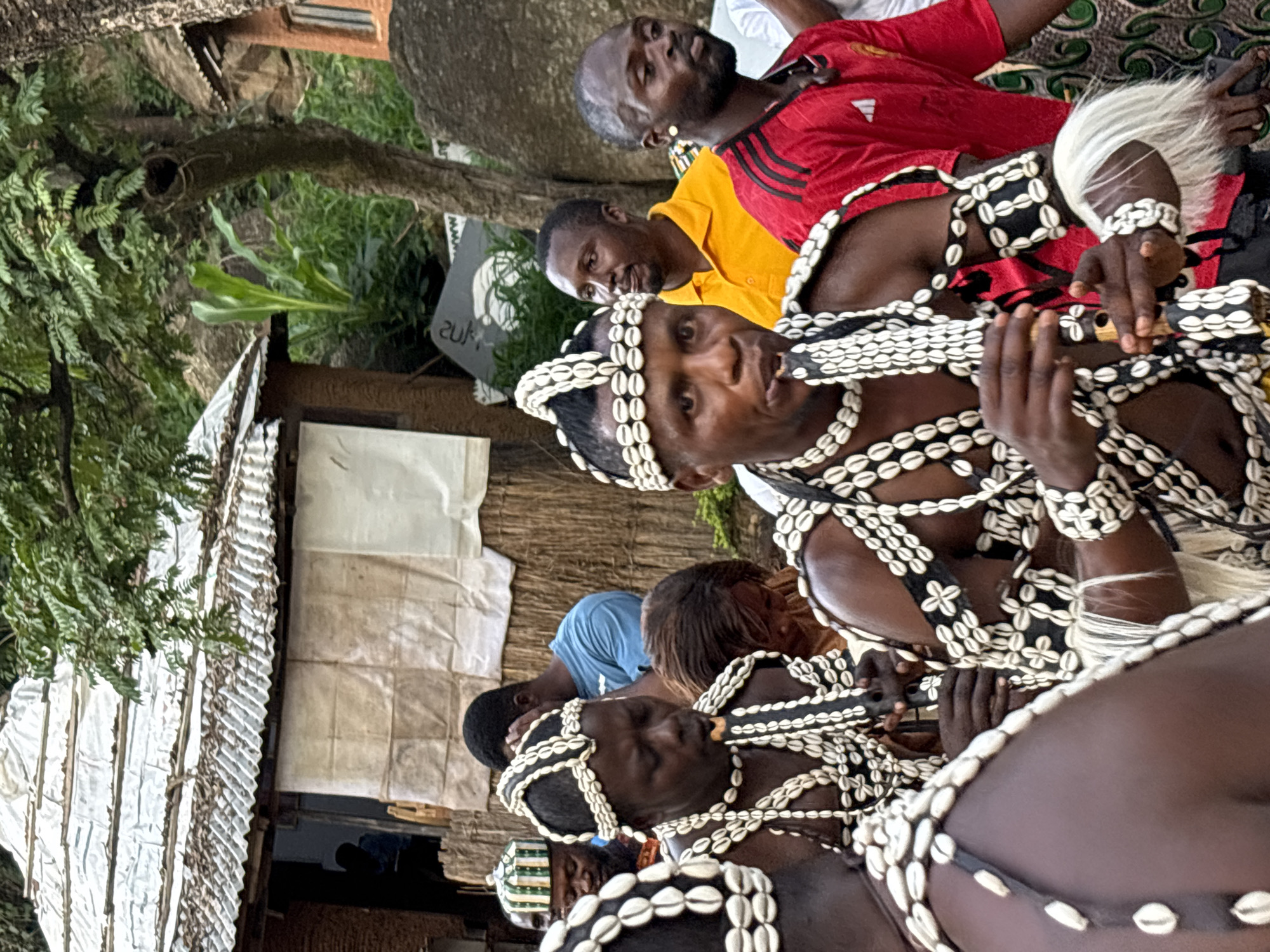 This image depicts a group of men in traditional attire, possibly part of a cultural ceremony or performance, in a village setting that appears to be in or around Jos, Nigeria. The central figures are adorned with elaborate headdresses and necklaces made of white shells. They are playing flutes, suggesting a musical element to the event.
The background reveals rustic structures with corrugated metal roofs and walls made of woven materials. Lush green vegetation and large rocks are visible, indicating a natural, possibly hilly, environment. The people in the background are dressed in a mix of casual and possibly ceremonial clothing, with one man wearing a bright orange shirt and another in a red shirt with black stripes. The overall atmosphere suggests a community gathering or a cultural event taking place outdoors.