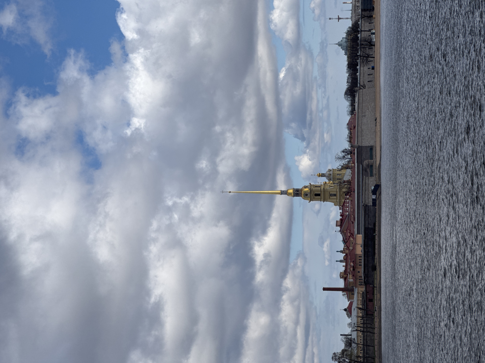 The image, oriented vertically, captures a panoramic view of the Neva River embankment in Saint Petersburg, Russia, with the iconic Peter and Paul Fortress in the distance. The most prominent subject is the golden spire of the Peter and Paul Cathedral, which stands tall against the sky in the middle ground, its golden dome visible beneath it.
In the foreground, a wide expanse of water, likely the Neva River, dominates the lower portion of the scene. The water surface is textured with numerous small ripples, indicating movement or a gentle breeze. Along the far bank, a line of buildings with red-tiled roofs stretches across the frame. Some bare trees and other structural elements are also visible along the waterfront.
The sky, filling the upper half of the image, features a dramatic display of cumulus clouds. Large, grey, and white cloud formations are interspersed with patches of clear blue sky, suggesting dynamic weather conditions on a daytime outing. The lighting is bright, consistent with daylight. No specific activities or people are visible, presenting a static yet impressive cityscape. There is no visible text in the image.