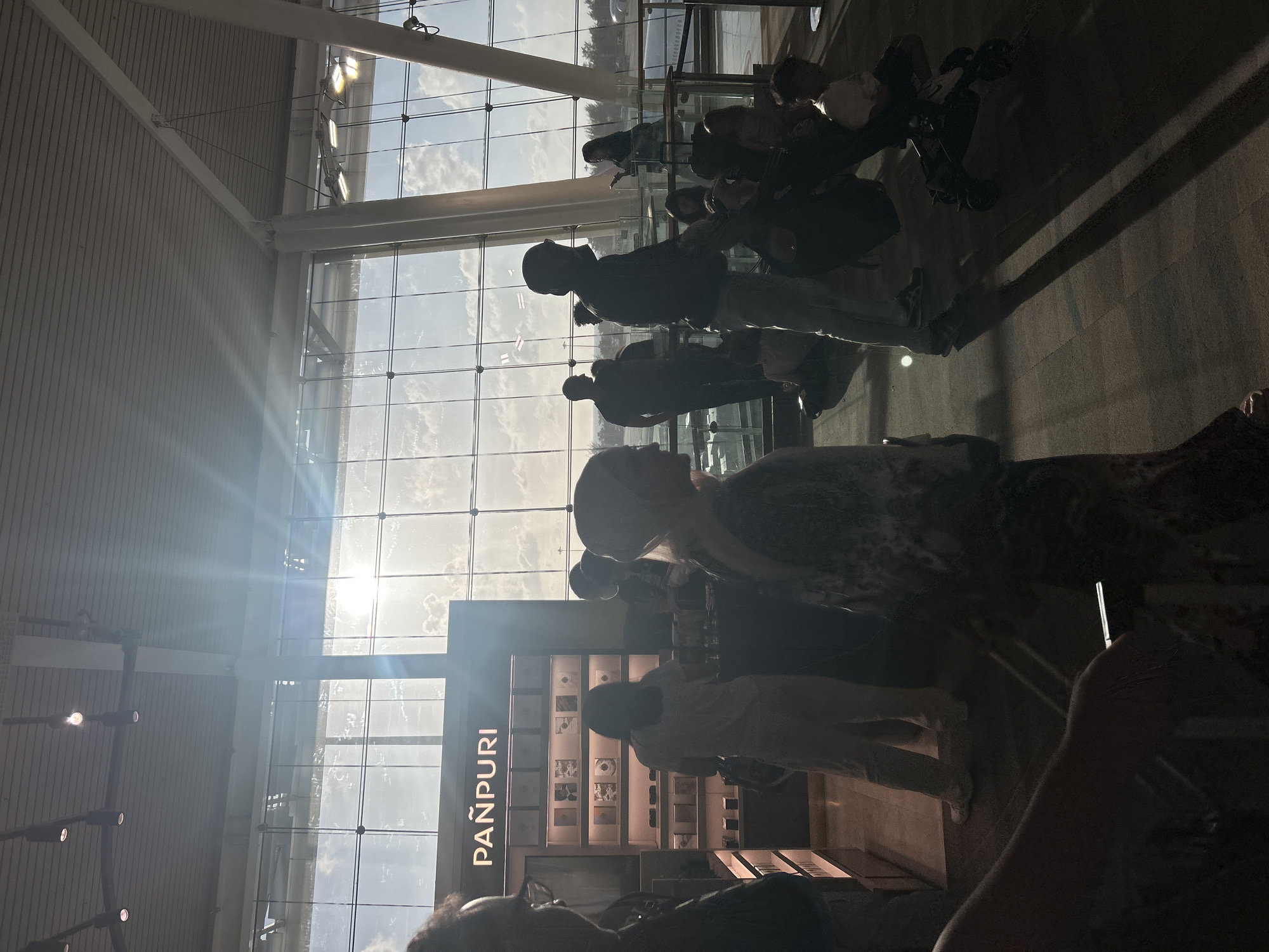 This image depicts an interior scene, likely an airport, with a prominent storefront displaying the brand "PANPURI." The storefront is illuminated and filled with shelves of products, suggesting it is a retail space. Large windows in the background reveal a bright sky with clouds, and an airplane wing is visible outside, further supporting the airport hypothesis. Several individuals are present, some are silhouetted against the bright windows, while others are in the foreground. One person appears to be interacting with the store's display. The lighting suggests it is daytime, possibly late afternoon due to the angle of the sun. The location is specified as Tambon Mai Khao, Thailand.