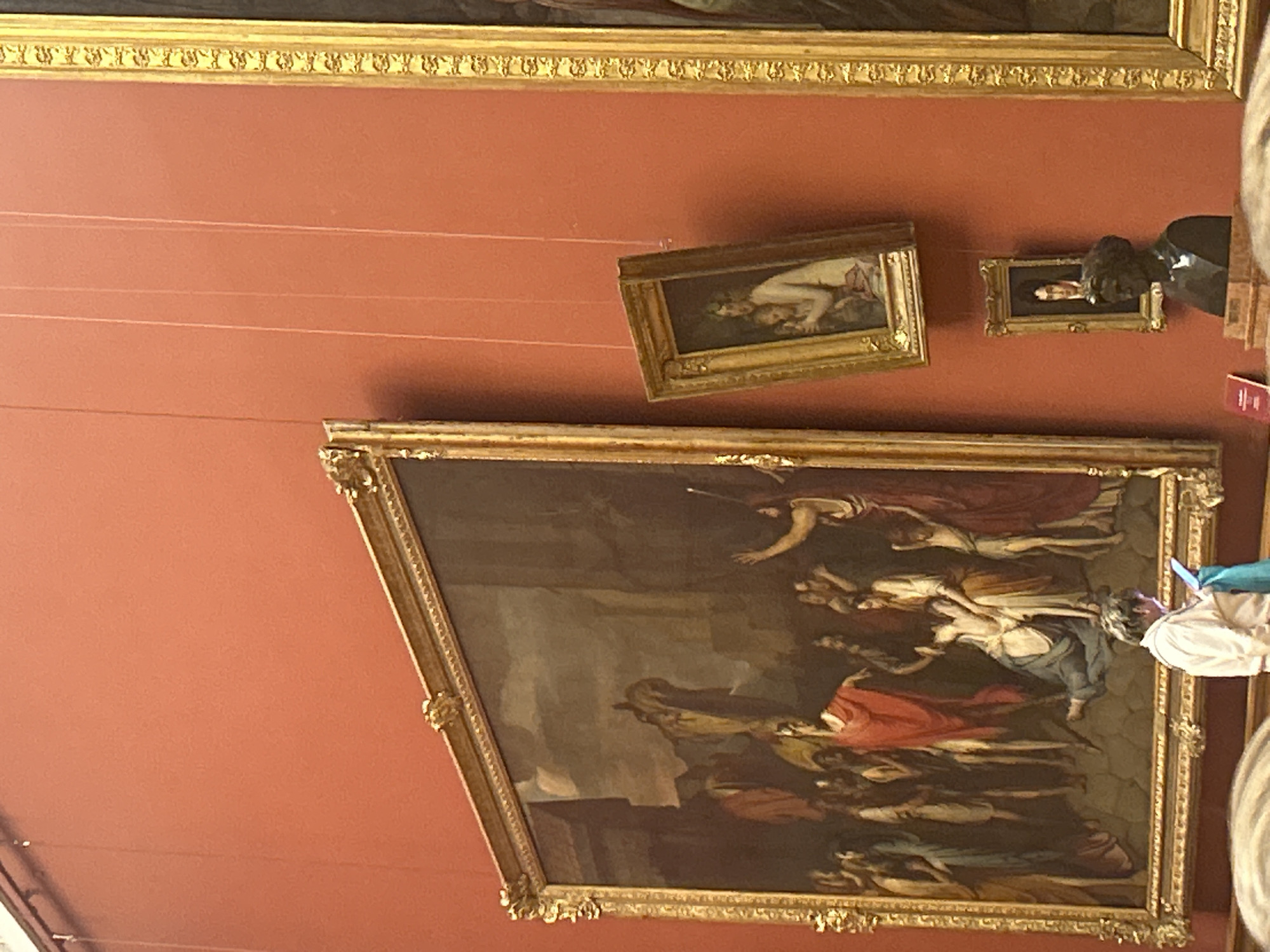 The image depicts an interior museum setting with several framed artworks displayed on a reddish-orange wall. Dominating the left side is a large, ornate, gilded frame enclosing a classical painting with numerous figures in draped clothing. To the right and slightly higher, a smaller, similarly framed painting is tilted, showing a woman with a child. Below this, another small framed portrait hangs, and further down and to the right, a bust of a man with dark hair is visible, resting on a marble pedestal. The scene is dimly lit, and the focus is on the artwork and the way it is presented, with the wall serving as a neutral backdrop. The overall impression is that of a formal exhibition space within an art museum. The specific location of this scene in Saint Petersburg, Russia cannot be determined from the image alone.