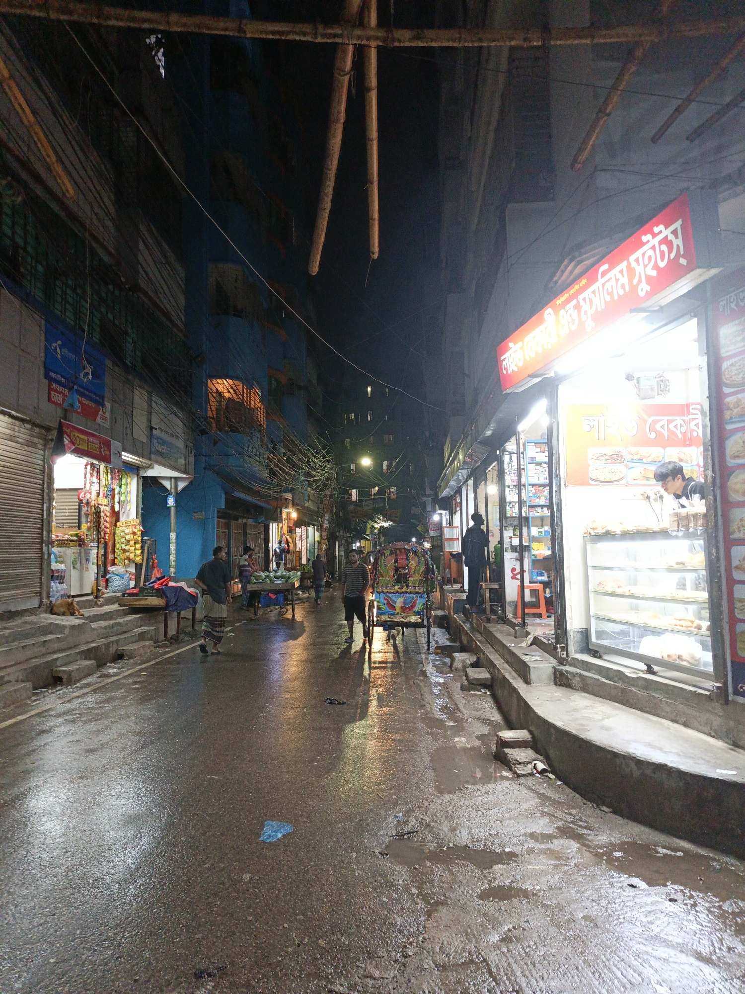 This is an image of a street in an unknown city in Unknown. The street is wet, likely from recent rain, and reflects the lights from storefronts and street lamps. A rickshaw with a passenger is in the middle of the street, and pedestrians are walking along the sidewalks. The storefronts are small and appear to sell food, with one being a bakery. Utility wires and bamboo scaffolding overhead create a dense network above the street. The buildings are multi-story and show signs of wear. The street is relatively narrow, and the lighting is dim except for the illuminated storefronts.