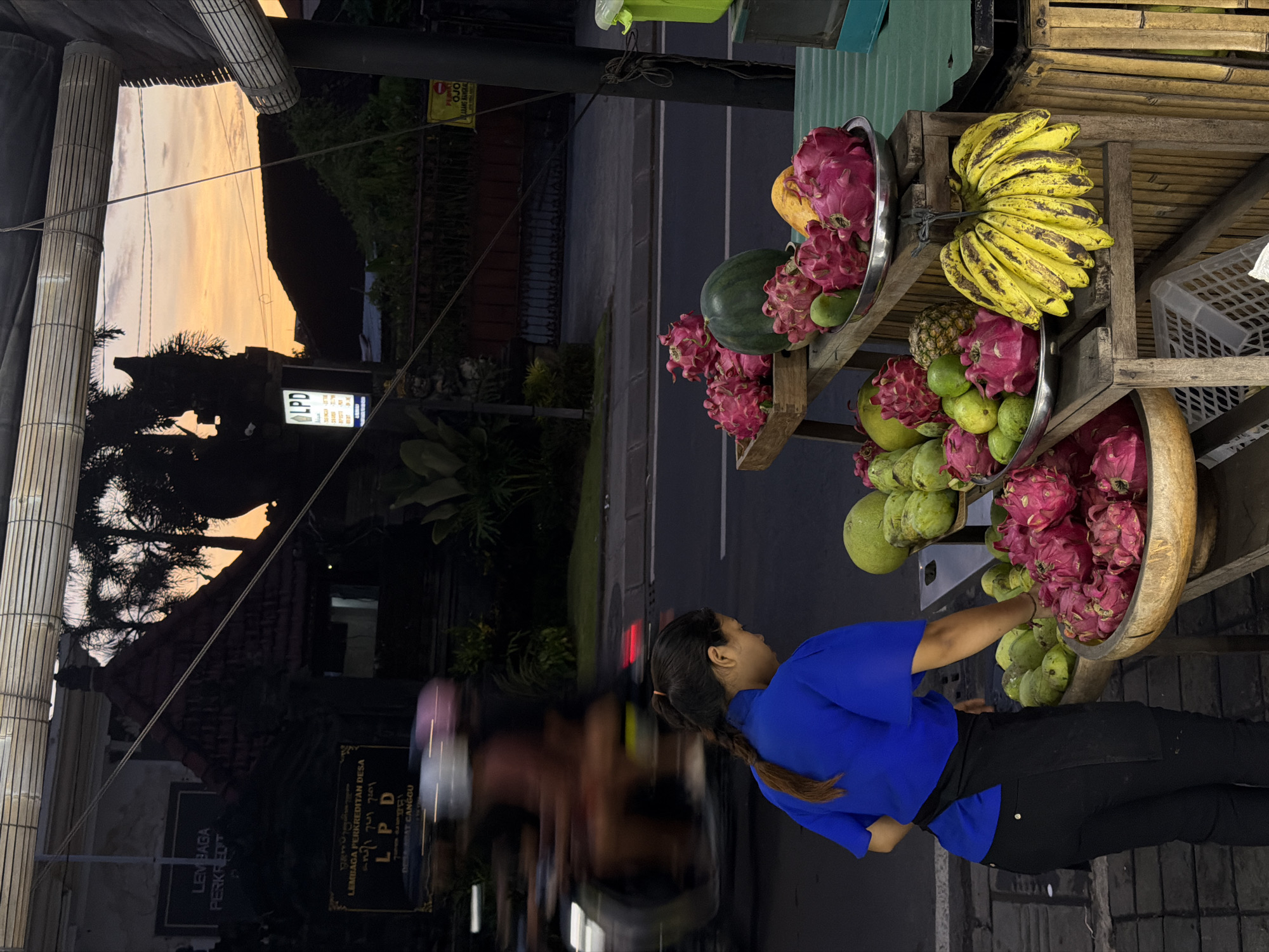 The image shows a woman reaching into a large wooden bowl filled with fruit, specifically dragon fruit and custard apples. This fruit stand is located outdoors, displaying a variety of produce, including bananas and watermelons, on a wooden rack with metal bowls. Across the street, a building features a sign that reads "Lembaga Perkreditan Desa" and "LPD", indicating a local institution in North Kuta, Indonesia, based on the text "DESA ADAT CANGGU" on the sign. A portion of the building is reflected in a dark, reflective surface, where the evening sky is also visible. The woman is wearing a blue shirt and black pants, and her hair is in a braid. A road and some vegetation can be seen behind the fruit stand.