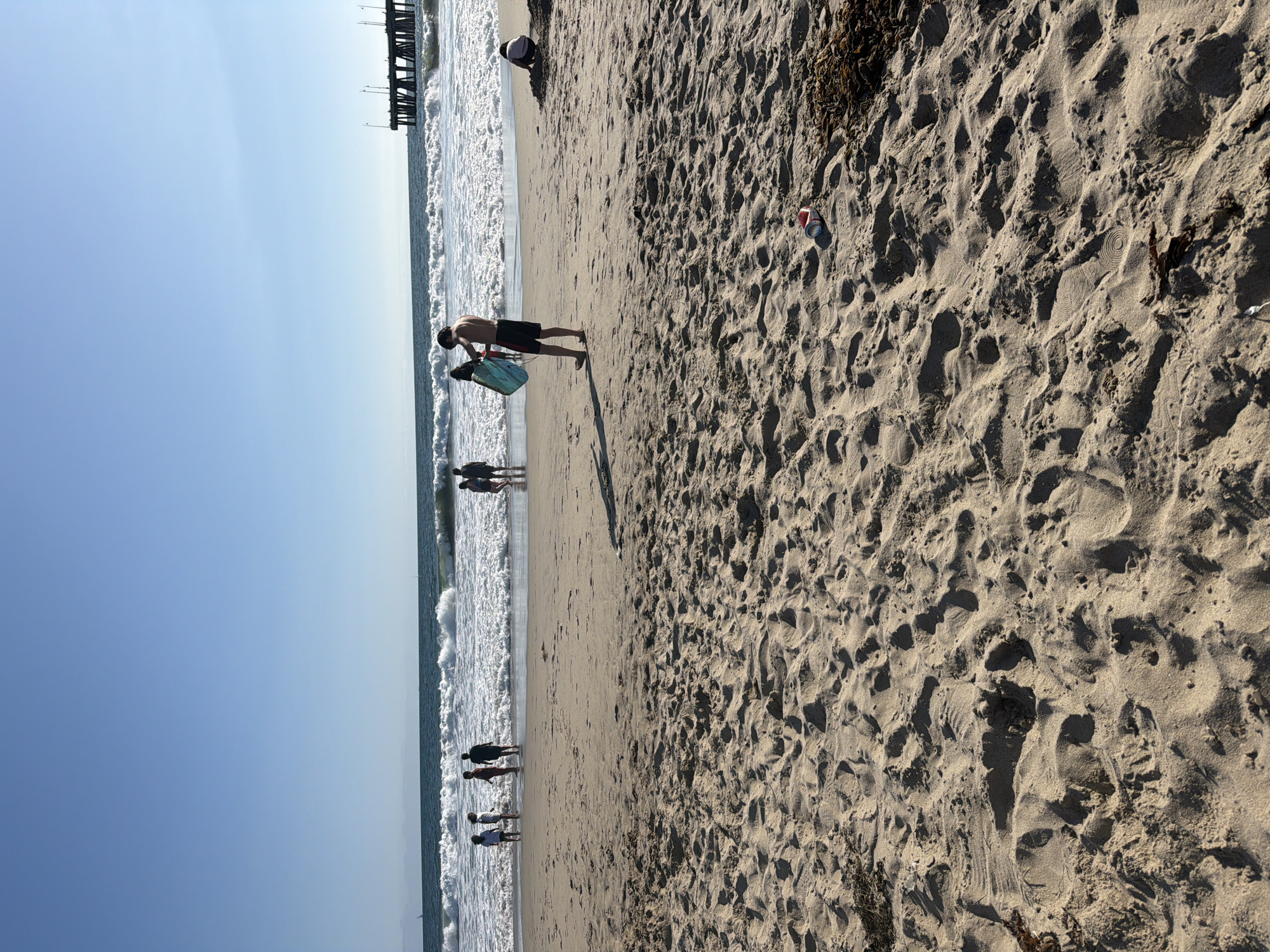A wide shot of a sandy beach shows several people enjoying the ocean and shoreline under a clear blue sky. In the foreground, the sand is textured with footprints and scattered debris, including a red and white can and seaweed. In the middle ground, a young man stands on the wet sand, holding a turquoise boogie board. To his left, a group of children are standing in shallow water. Further back, a long pier extends into the ocean, with seagulls perched on its structure. To the right, a person is sitting in the sand. The waves are breaking on the shore, creating white foam.