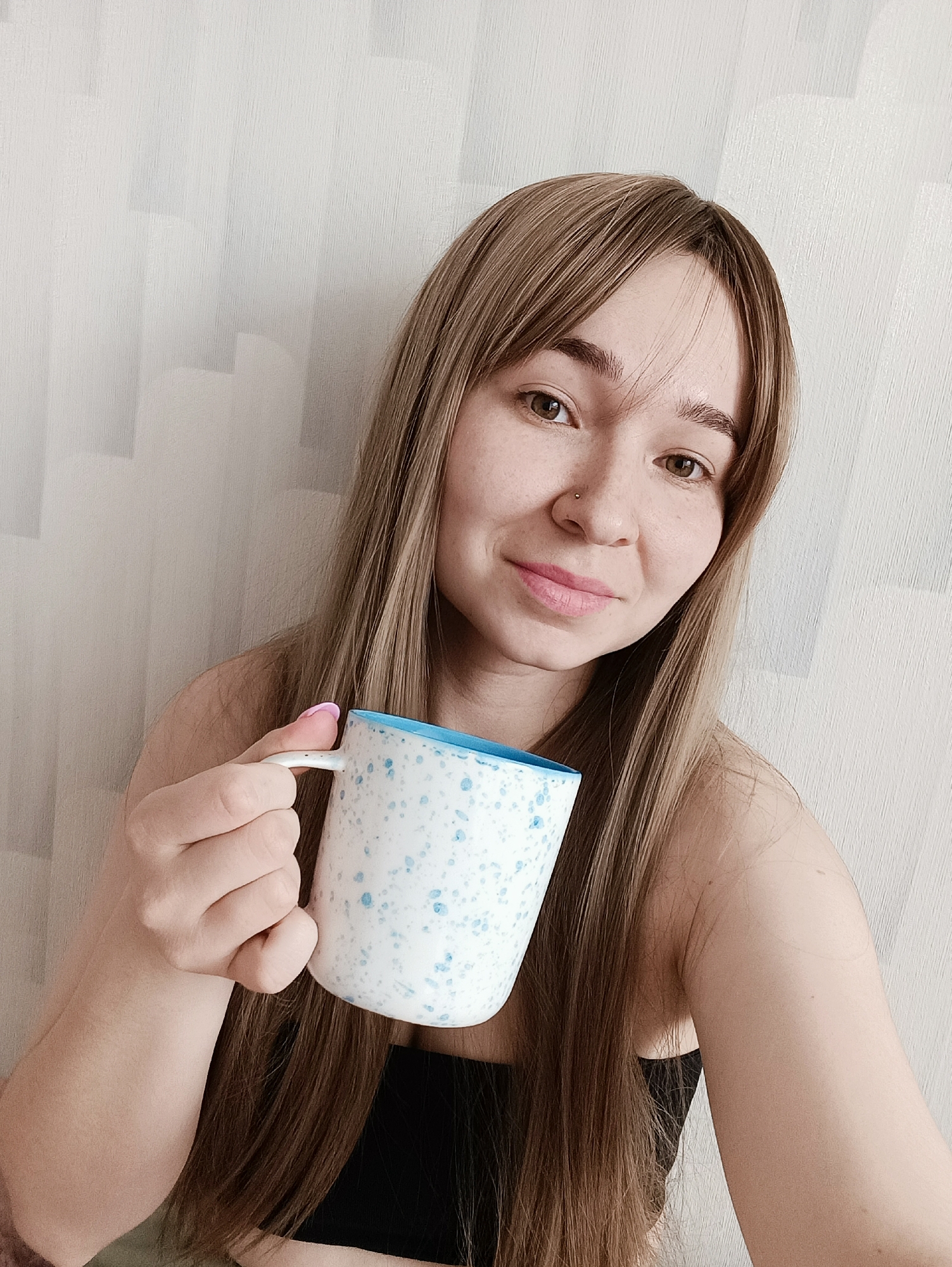 A woman with light brown hair and fair skin is holding a white mug with blue speckles and a blue interior. She is smiling slightly and looking at the camera. She appears to have a nose piercing. The background is a white wall with a subtle textured pattern.
There is no information to support that this picture was taken in Izhevsk, Russia.