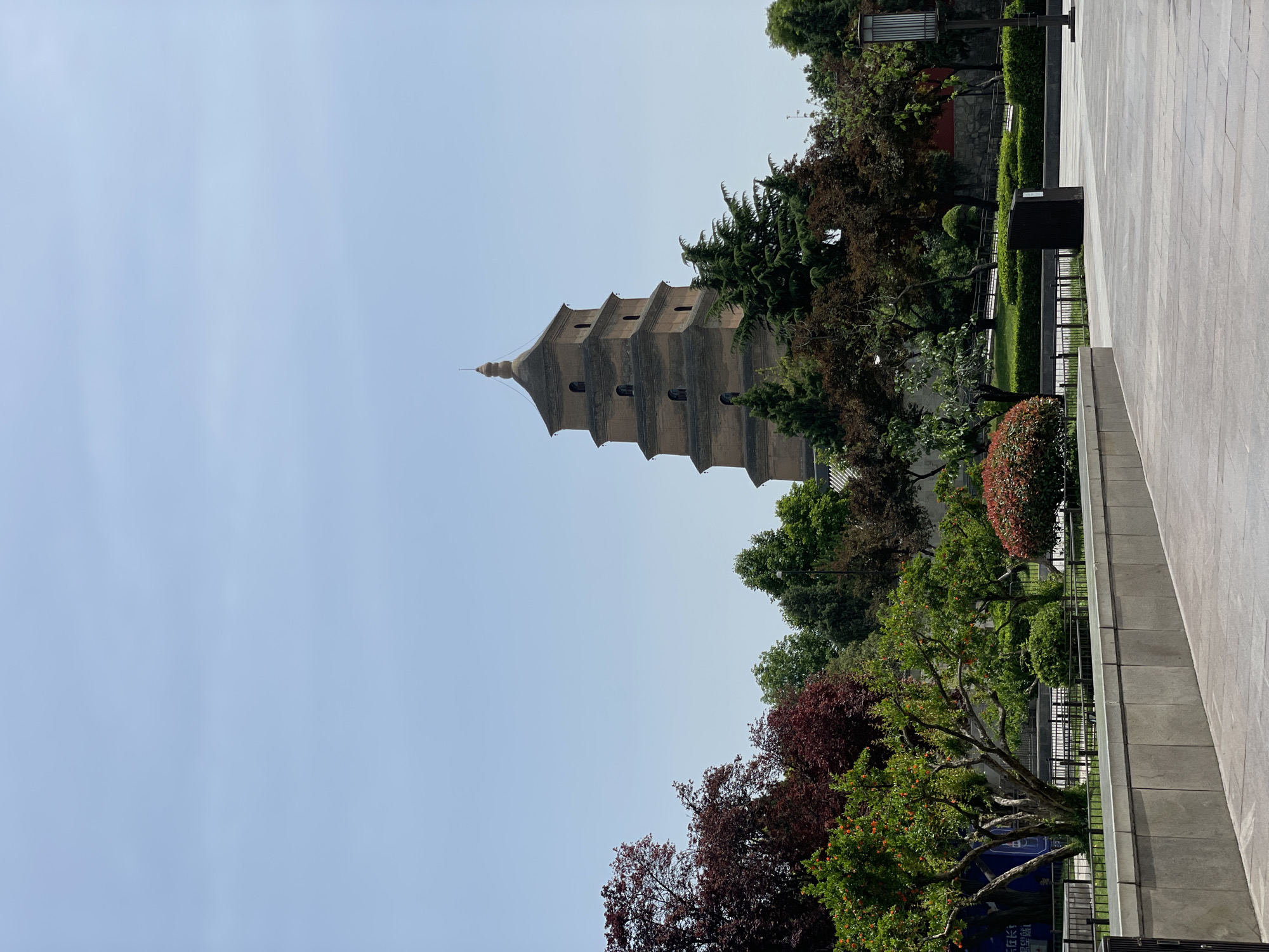 The image shows the Giant Wild Goose Pagoda in Yanta District, Xi'an, China. The pagoda, a multi-tiered, brick structure, is partially obscured by trees. The foreground shows a paved area and a railing with vegetation behind it. The sky is pale blue.