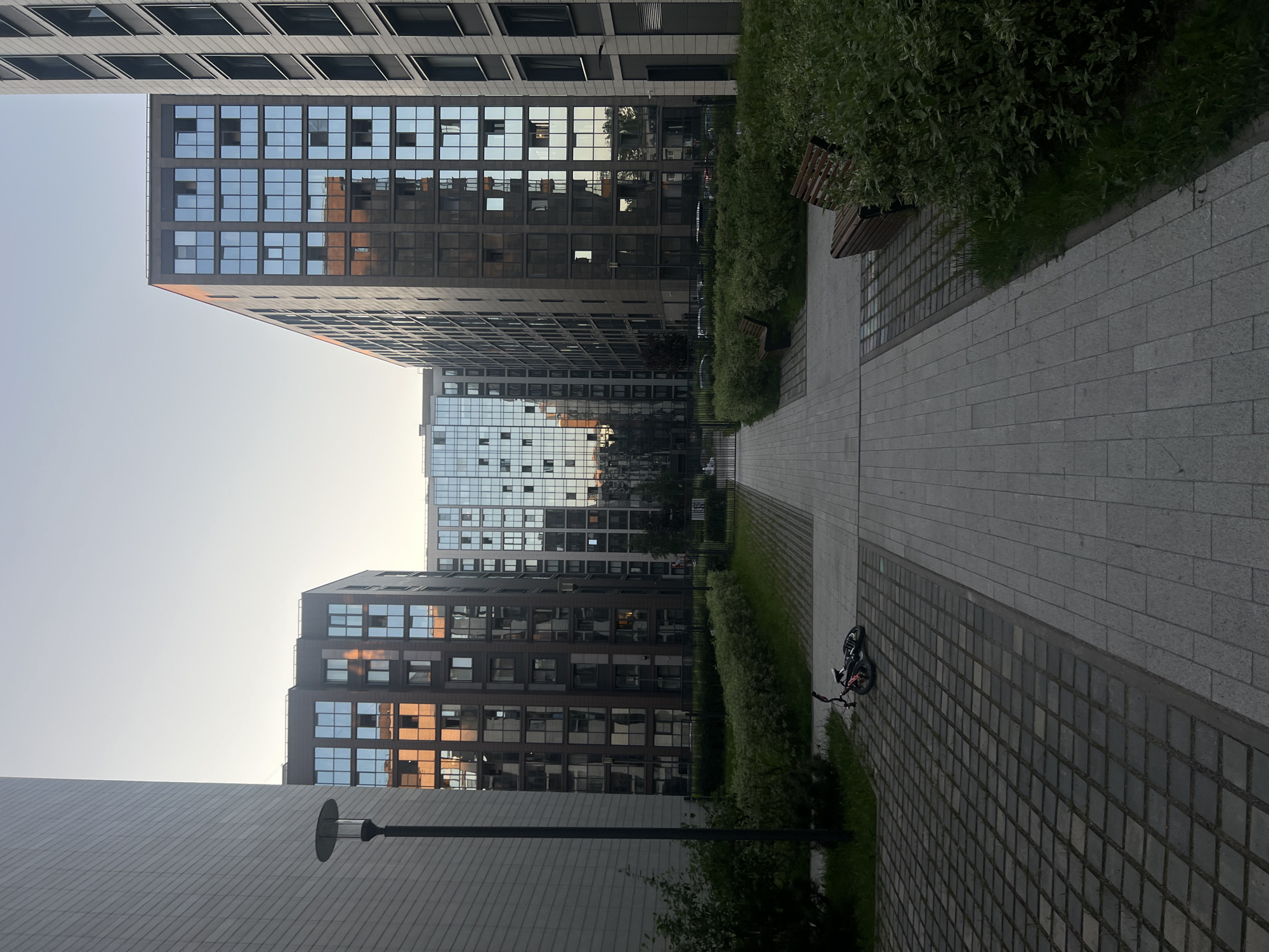 This image captures a modern urban courtyard or pedestrian zone, located in the city of Unknown, Unknown, during either early morning or late afternoon twilight. A light grey paved walkway forms a central axis, receding into the background between two multi-story buildings. This pathway is flanked by darker grey square-patterned pavers and neatly trimmed green hedges.
On the left side of the frame, a large, light grey building with horizontal paneling rises vertically, partially obscuring the scene. A dark, slender lamppost with a circular top is positioned in front of this building. To the right, two prominent buildings with dark grey or brown facades feature numerous reflective glass windows, exhibiting strong orange and golden reflections from an external light source, likely the setting or rising sun. A lighter-colored building with similar glass architecture is visible at the end of the central walkway in the distance.
On the dark grey pavers to the left of the central path, a small black and red bicycle lies on its side. On the right side, two dark wooden benches with metal frames are partially visible, set back behind the green hedges. A black metal fence runs horizontally across the mid-ground, separating the landscaped area from structures further back. The sky above is clear and pale blue, indicating fair weather. No individuals are present in the scene.