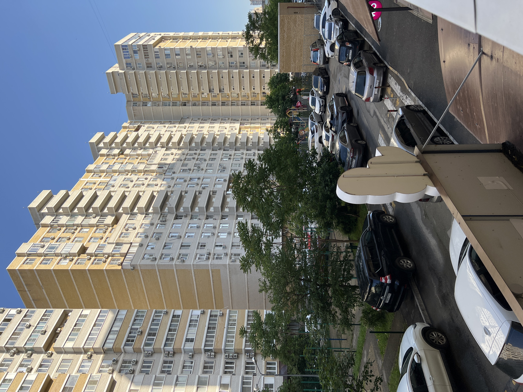 The image shows a street scene in Krasnodar, Russia. Several multi-story residential buildings are visible, predominantly constructed with light-colored exteriors and many windows. A line of parked cars fills the right side of the frame along a narrow street. Trees are interspersed among the buildings and cars. A playground can be seen in a small space between the buildings. The sky is clear and blue.