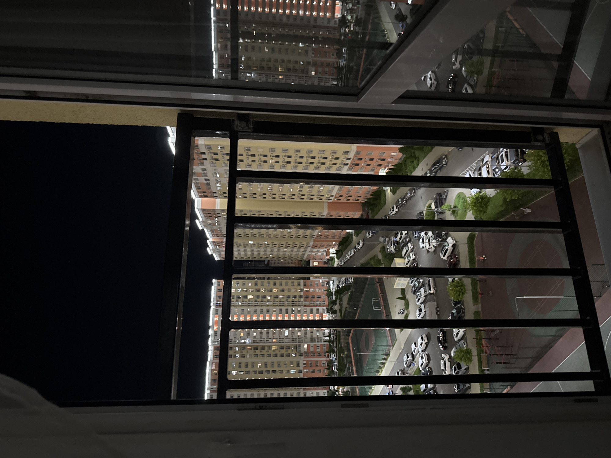 The image captures a nighttime view from a balcony overlooking a residential complex in Anapa, Russia. Two large, multi-story apartment buildings, illuminated by numerous windows, dominate the scene. The buildings are modern with a color scheme of beige, orange, and white. In front of the buildings, there is a sports court, possibly a tennis court or basketball court, with red and green surfaces. Adjacent to the court is a parking lot filled with various cars, indicating residential activity. The overall environment suggests a well-developed urban area at night, with artificial lighting illuminating the buildings and surrounding infrastructure.