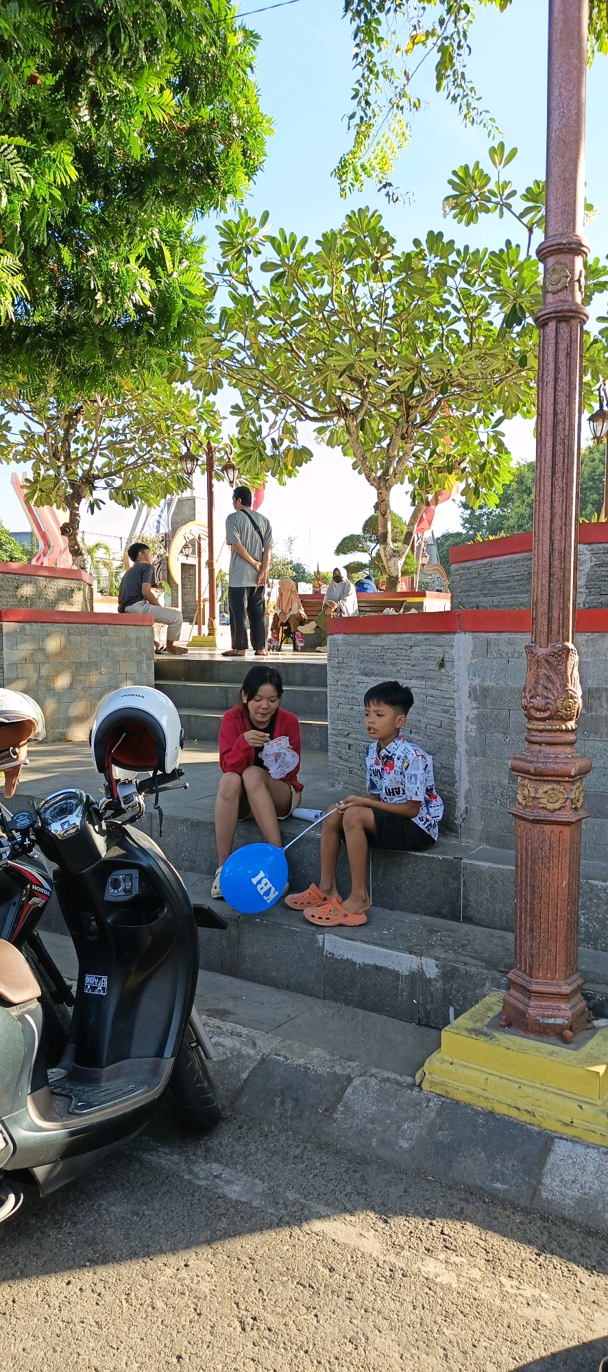 In Klaten, Indonesia, two children, a girl wearing a red shirt and shorts, and a boy in a patterned shirt and black shorts, are sitting on concrete steps. The girl is holding a crumpled plastic bag, and the boy is holding a blue balloon with "KBI" written on it. In the foreground to the left, a black scooter is parked, with a white helmet on its handlebars. Behind the children, other people are visible sitting on benches or standing. The scene is outdoors, with trees providing shade, and the sky is clear blue. The steps and surrounding structures appear to be made of stone and concrete, with decorative red trim. A lamppost with a weathered, ornate metal design stands to the right of the frame.