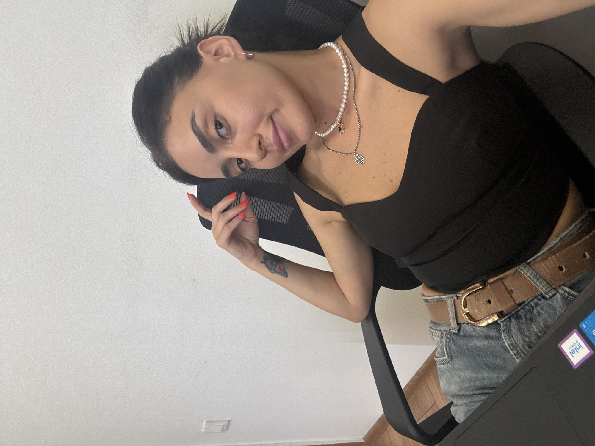 A young woman with dark hair and brown eyes, wearing a black tank top, blue jeans, and a brown belt, is seated in a black office chair. She has a pearl necklace with two pendants, one a dark gem and the other a silver cross. A tattoo of a butterfly is visible on her left arm. Her right arm is resting on the back of the chair, and her left hand is near her face. A laptop with an "Intel" sticker is in front of her. The scene is indoors, with a white wall behind her. There is no specific indication of the location being in Tbilisi, Georgia.