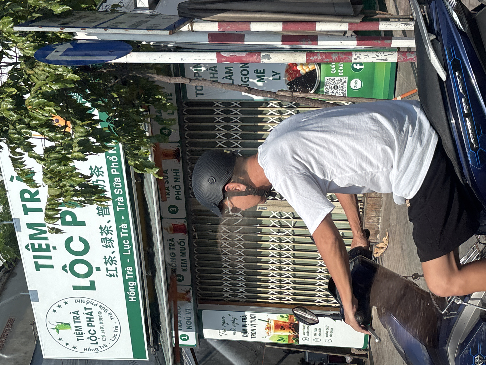 The image shows a person on a motorcycle in front of a tea shop. The shop has a large sign that says "TIỆM TRÀ LỘC PHÁT" in Vietnamese, and lists various types of tea, including "Hồng Trà - Lục Trà - Trà Sữa Phở Mì". The shop appears to be located in Nha Trang, Vietnam, as indicated by text on the sign and a visible QR code associated with "Zalo", a popular messaging app in Vietnam. The person on the motorcycle is wearing a white t-shirt, black shorts, and a black helmet. The motorcycle is blue and black. In the background, there are more signs for the tea shop and a street with buildings and trees. The overall scene depicts a typical street view in a Vietnamese city.