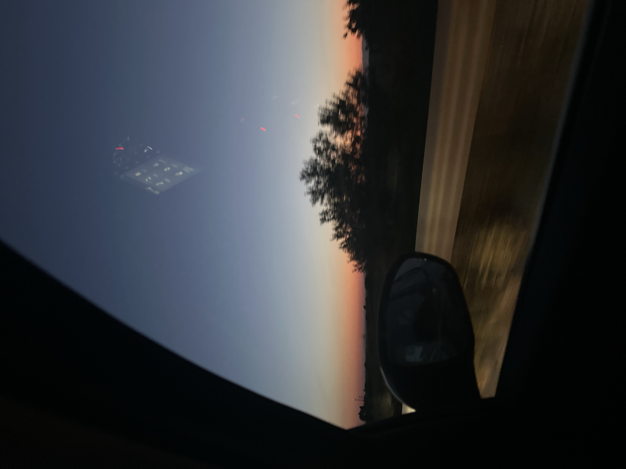 The image depicts a view from inside a vehicle at dusk, looking out the side window towards a twilight sky. A car's side mirror is visible in the foreground, reflecting the road and trees. The sky transitions from a deep blue at the top to warm orange and pink hues near the horizon, indicating sunset. Silhouetted trees line the horizon, with some appearing blurred due to motion. A faint reflection of a building with illuminated windows can be seen in the upper portion of the sky. The road surface, visible through the window, is illuminated by streetlights or headlights, creating a streaky, golden effect. The overall impression is one of a journey through the countryside at the end of the day. There are no people visible in the image. The location is stated to be the city of Skutare, Bulgaria.