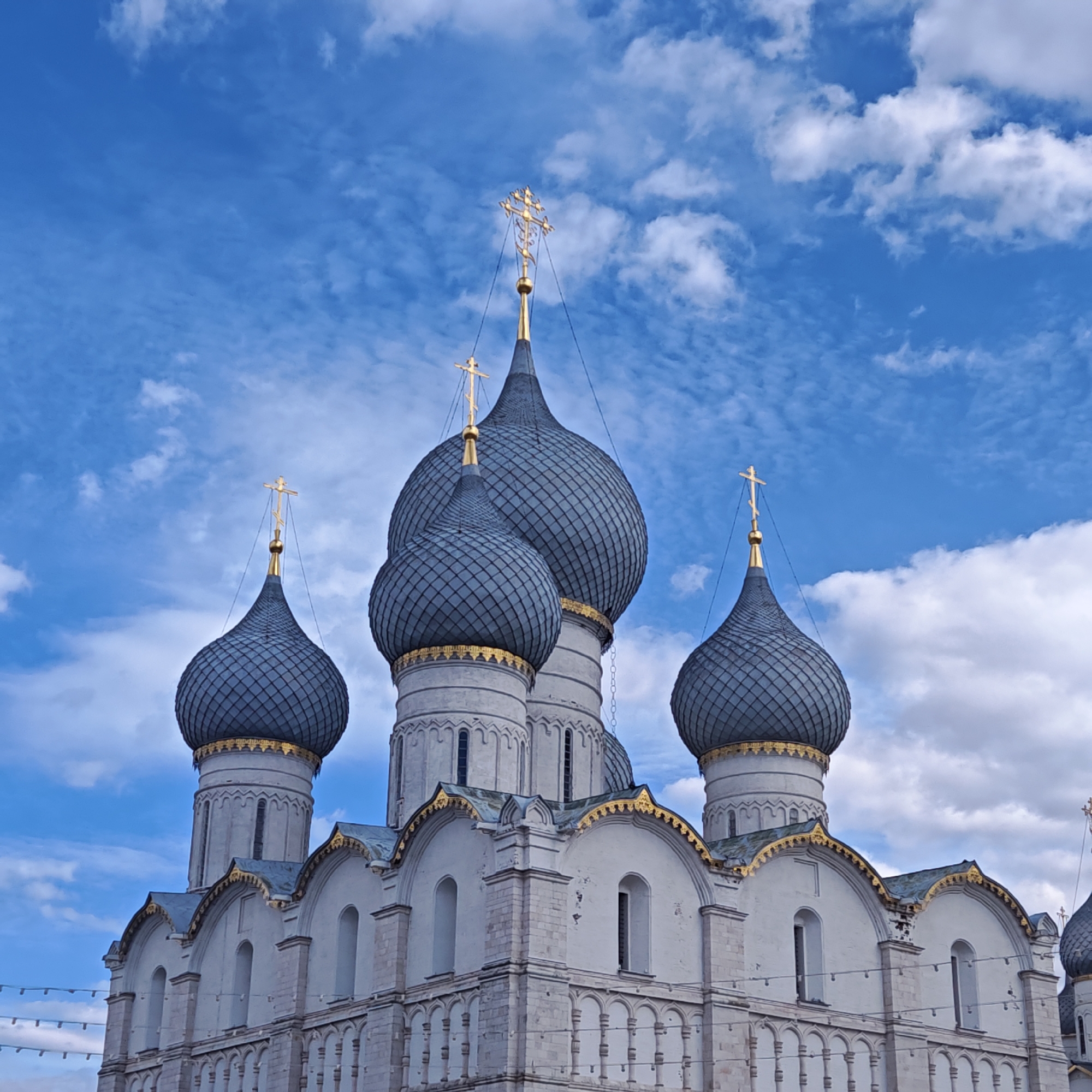 This image shows the Dormition Cathedral in Rostov, Russia, from a low angle. The cathedral features white walls and five onion-shaped domes covered in gray scales with gold accents. Each dome is topped with a gold cross. The sky is blue with scattered white clouds. There are decorations hanging on the bottom of the wall.