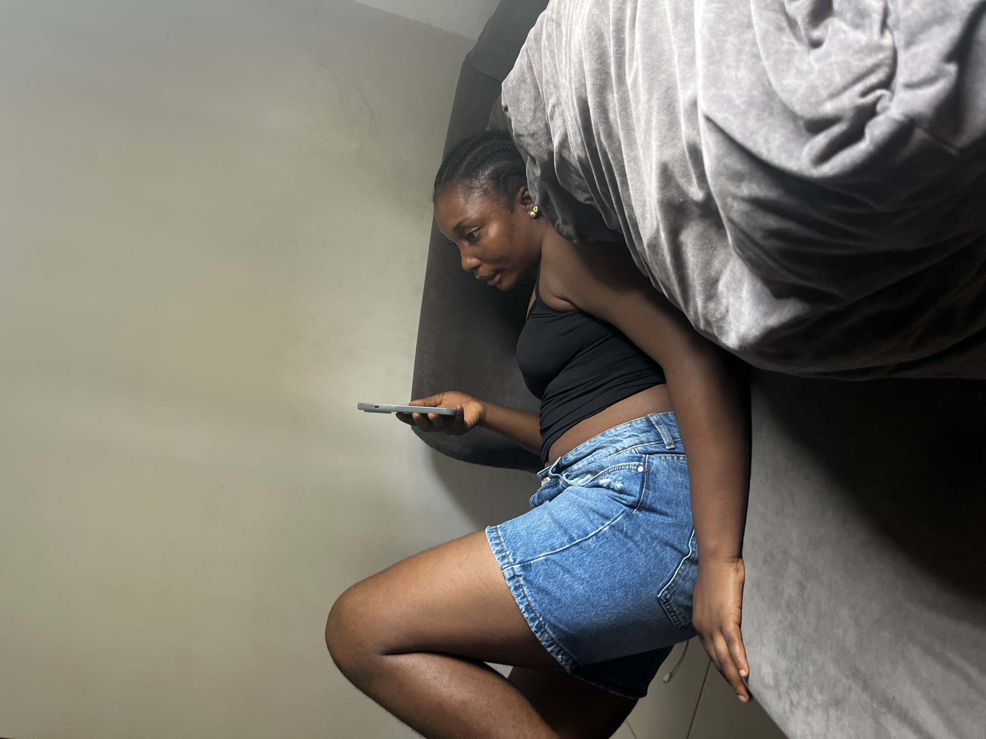 A young Black woman is lying on her side on a grey sofa, propped up against a wall. She is wearing a black tank top and denim shorts. Her hair is braided in cornrows. In her right hand, she holds a grey smartphone, looking down at it. Her left arm is bent and resting on the sofa cushion. A grey blanket is draped over her legs and the sofa. The background is a plain, cream-colored wall. The lighting suggests an indoor setting, possibly a living room.