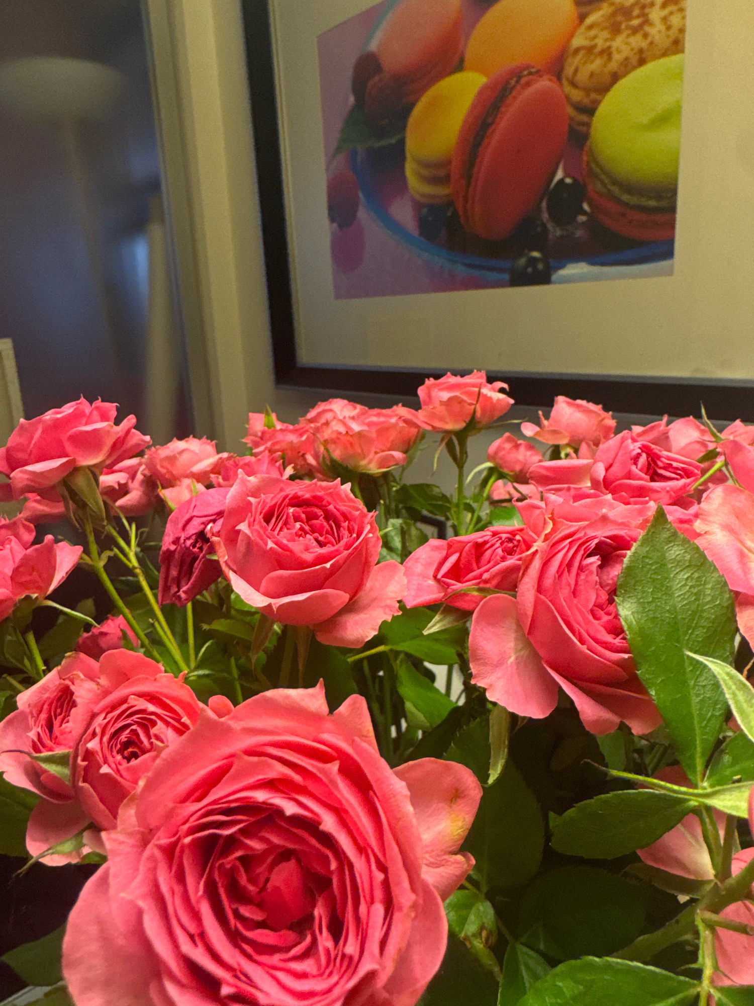 A close-up shot of a bouquet of pink roses with green leaves is in the foreground. In the background, a framed picture of colorful macarons is visible on a wall. The scene appears to be indoors, possibly in a home or a shop. There is no indication of the location being in Almaty, Kazakhstan. The image is well-lit and the focus is on the roses.