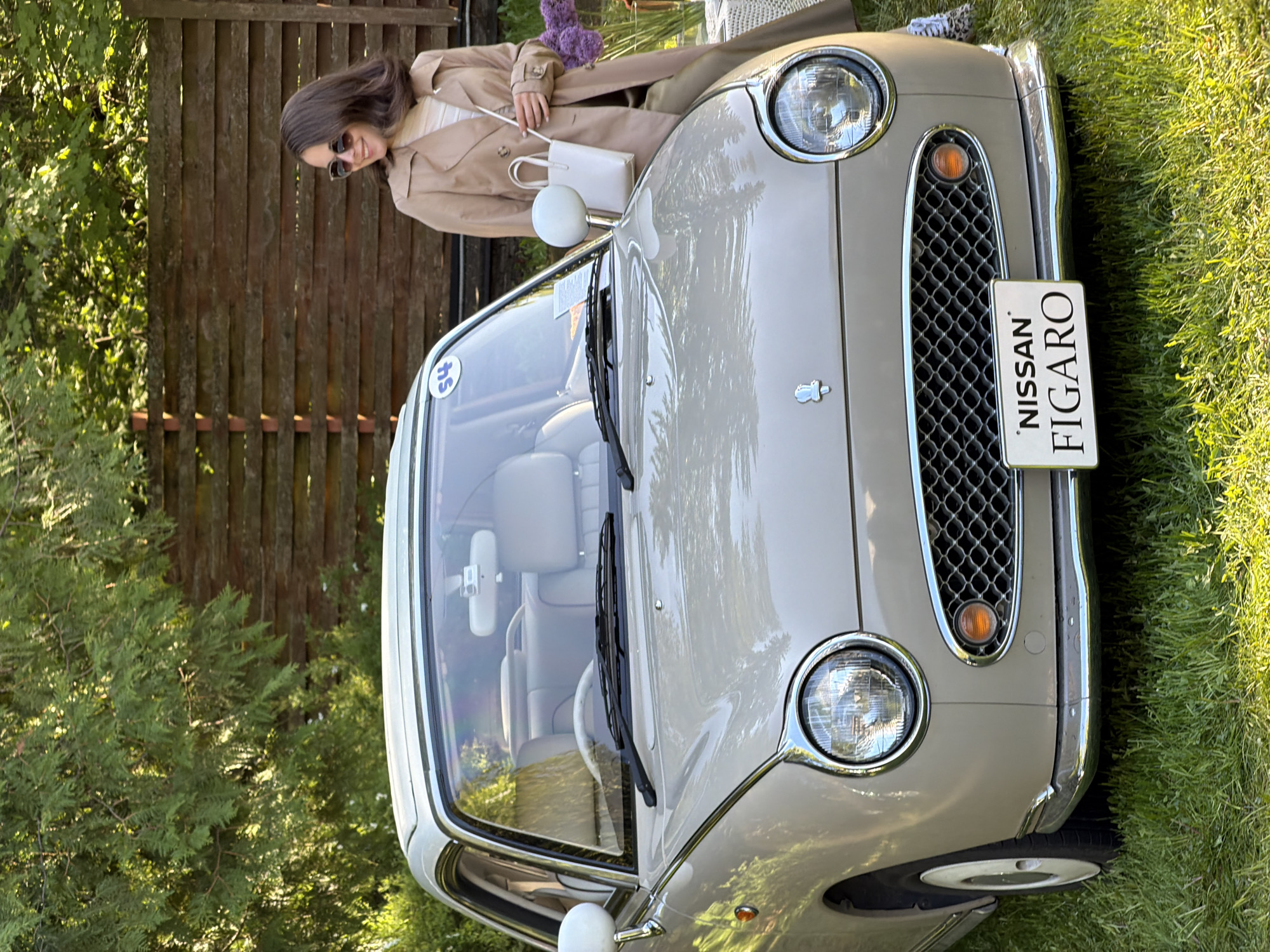 The image features a tan Nissan Figaro automobile parked on a lawn with a woman posing next to it, potentially in Repino, Russia. The car, characterized by its retro design, is positioned in the foreground. The "NISSAN FIGARO" license plate is clearly visible. The woman is dressed in a beige trench coat and sunglasses, standing partially on the hood of the car, holding a small bouquet of purple flowers and a white purse. A wooden fence is visible in the background, partially obscured by greenery and a tree on the left side. The overall environment is outdoors, suggesting a sunny day.