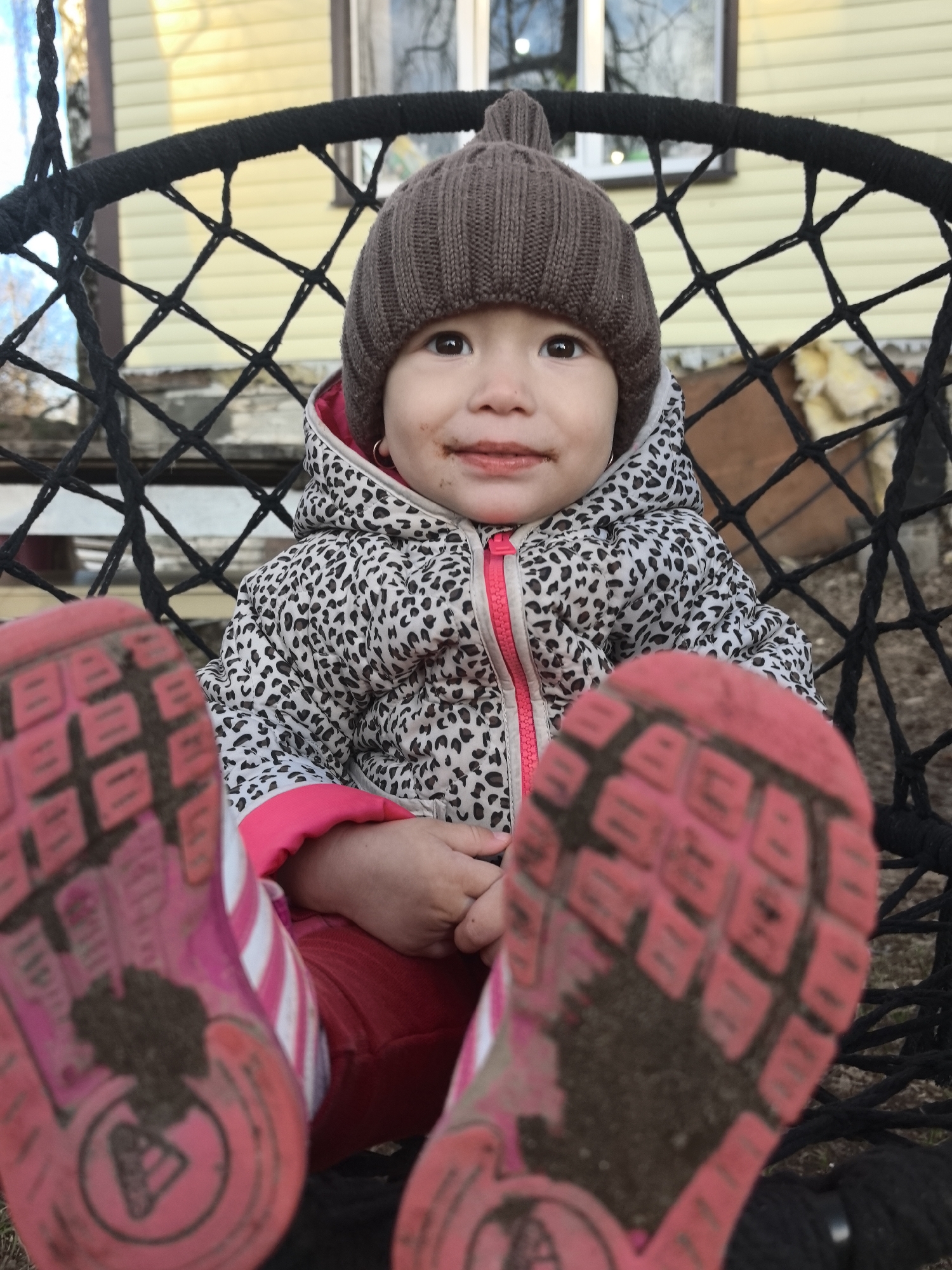 A young child sits in a black, woven hanging chair with a yellow building in the background. The child wears a brown knit hat, a jacket with a white and black print and pink zipper, red pants, and pink sneakers with white stripes. The soles of the sneakers are visible and covered in dirt. The child appears to have dirt or chocolate around the mouth.