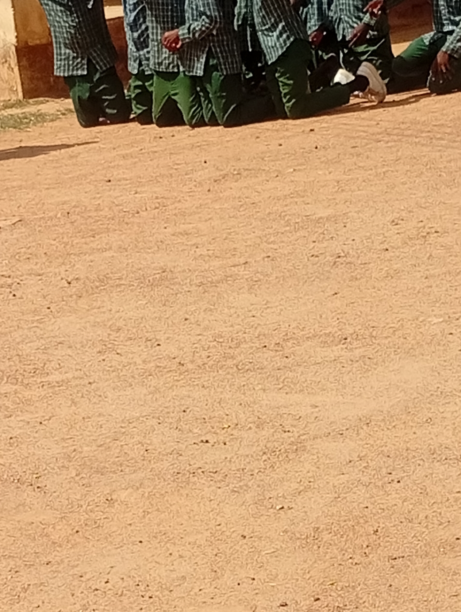 The image shows the legs and torsos of several individuals, likely students, kneeling on a sandy or dirt ground. They are wearing plaid shirts and green trousers. One person on the right is wearing white sneakers, while another on the left is wearing black shoes. The background on the left shows a portion of a building, possibly made of mud bricks or concrete. The scene appears to be outdoors, in a dusty or sandy area. The location is identified as Dutse, Nigeria.
