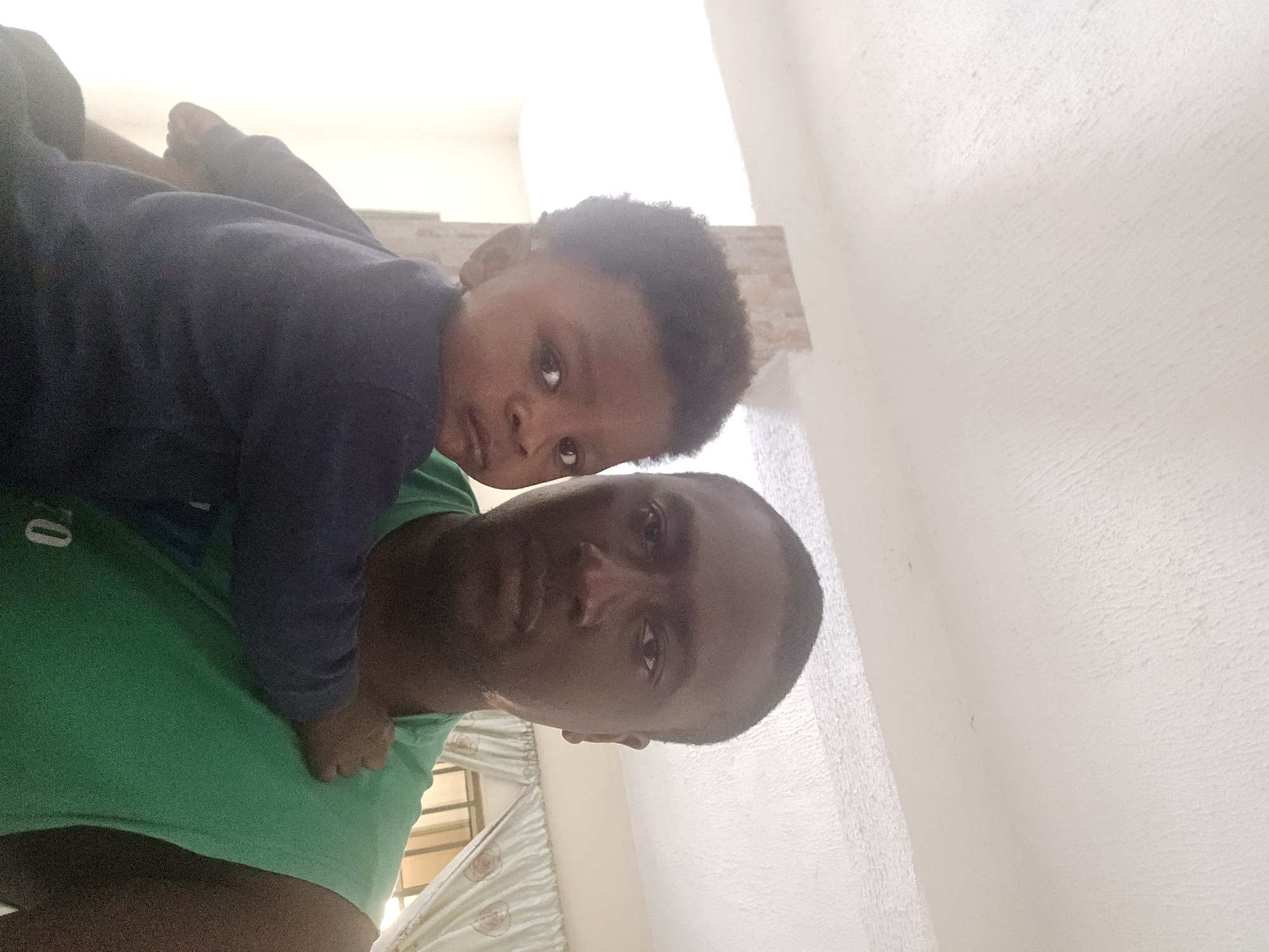 In this selfie-style photograph, a Black man, likely in his late 20s or early 30s, holds a young Black child, approximately one to two years old, in his arms. The man is wearing a sleeveless green t-shirt and is looking directly at the camera with a neutral expression. The child, with curly dark hair, is positioned in front of the man, facing the camera and looking slightly to the left with a curious expression. The child is wearing a dark blue long-sleeved shirt.
The background suggests an indoor setting with a lightly textured, off-white ceiling that appears to be illuminated by ambient light. To the right of the subjects, sheer, light-colored curtains with a subtle pattern are visible, partially obscuring a window or another room. A portion of a vertical, textured architectural element, possibly a column or a wall, is visible behind the child on the left. The lighting in the room is bright, casting some soft shadows and highlights on the subjects and the environment. The overall impression is a casual, domestic moment.
While the image itself does not contain explicit markers of location within Jos, Nigeria, the context provided suggests it was taken there.