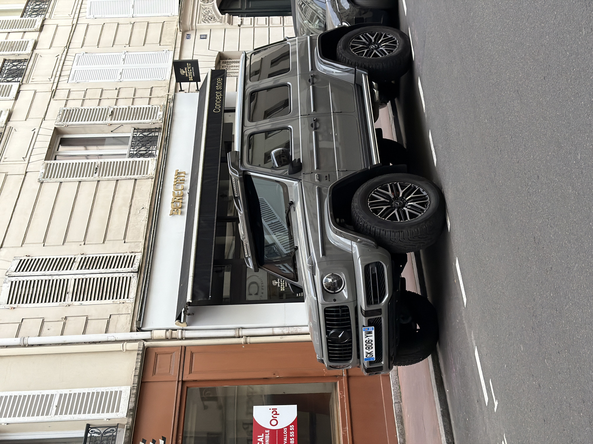 A grey Mercedes-Benz G-Wagen SUV is parked on a paved street next to a multi-story building. The vehicle, visible from the front and side, features large, dark alloy wheels and a distinctive grille. Its French license plate, "GK-806-YM," is clearly visible on the front.
The building displays classic Parisian architectural elements, including light-colored stone or stucco facades, multiple windows with white louvered shutters, and decorative wrought-iron window guards on some lower floors. A black awning extends from a ground-floor shop, prominently displaying "BERECHIT Concept store" in golden lettering. A smaller black sign above the awning also shows "BERECHIT." The building's facade bears the number "11."
Adjacent to the "BERECHIT" store, another ground-floor unit features a wooden-framed storefront. A red and white real estate sign, partially obscured but legible, reads "LOCAL DISPONIBLE," "Orpi VALLOIS," and includes the phone number "01 41 05 55 55." This sign, particularly "VALLOIS," confirms the location context of Levallois-Perret, France.
The street is asphalt with white parking lines. The scene appears to be set during daytime, with soft, diffused lighting suggesting an overcast sky. There are no people visible in the image.