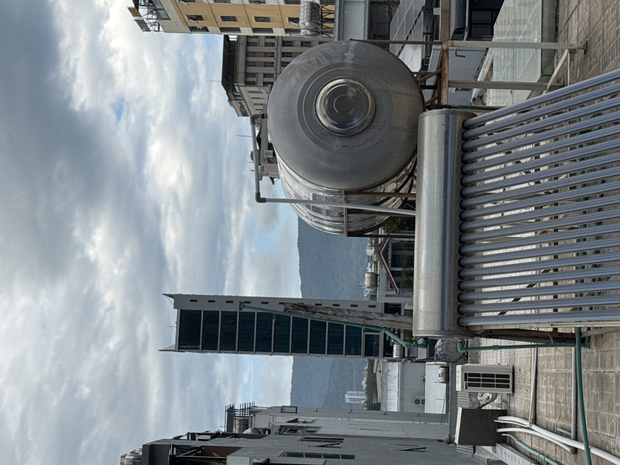 This is a rooftop view in Da Nang, Vietnam, on a cloudy day. In the foreground, a solar water heater with many blue tubes is prominently displayed. Behind it, a large, silver cylindrical water tank is mounted on a metal frame. Various pipes and plumbing are visible on the rooftop, connecting to the solar heater and the tank. To the left, a grey building with windows and an air conditioning unit is partially visible. In the mid-ground, several other buildings of varying heights and architectural styles are scattered across the urban landscape. Further in the background, lush green mountains rise under the overcast sky. The overall scene depicts a typical urban rooftop environment in Da Nang, equipped for water heating and energy collection, with a backdrop of the city and its natural surroundings.