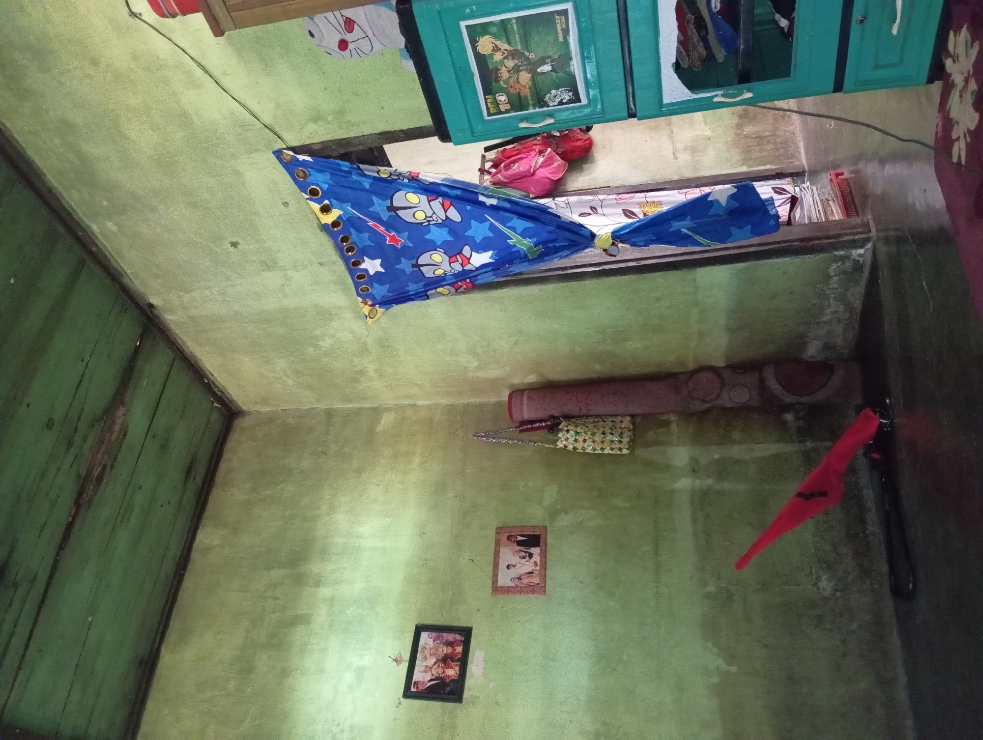 The image shows the interior of a room with green-painted walls and a green-painted wooden ceiling. In the upper left corner, a framed photograph of a couple in wedding attire is visible on the wall. Below it, another framed photograph displays a family group. To the right of the photographs, a rolled-up mat is leaning against the wall, and a woven bag is hanging next to it. A bright red object, possibly a tool or toy, lies on the floor in the lower left corner.
Further to the right, a blue curtain adorned with white stars and cartoon characters is drawn across a doorway. Behind the curtain, a pink backpack and a red bag are visible. A green plastic cabinet with drawers stands to the right of the doorway. The top drawer of the cabinet features a picture from the animated series "Ben 10." The room has a tiled or concrete floor, which is dark and reflective. The overall impression is of a simple, lived-in space. The mention of Rejosari, Indonesia, suggests a domestic setting in that location, likely a residence.