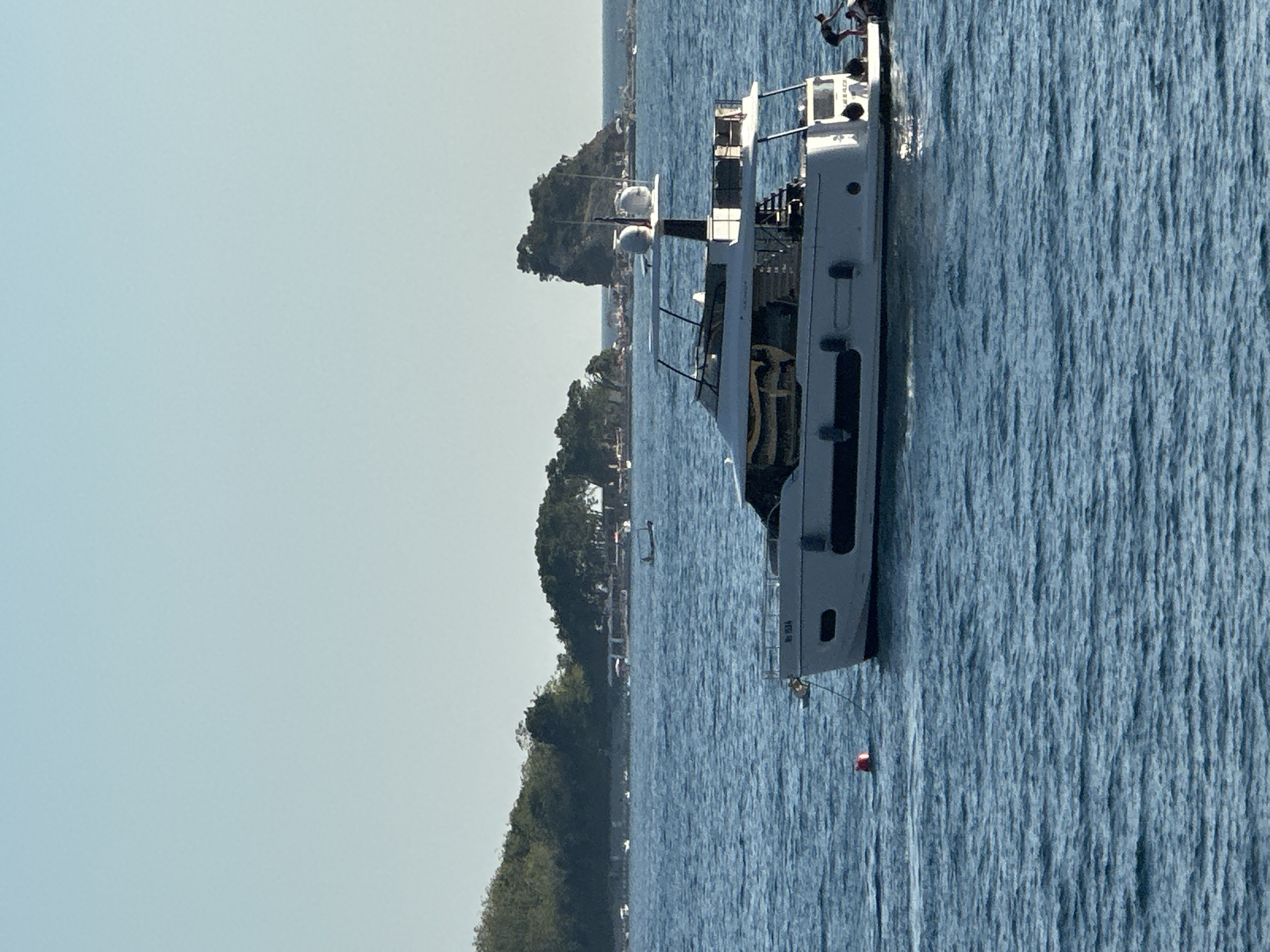 An elevated, eye-level, medium shot captures a white yacht sailing on a tranquil, blue sea under a clear, light blue sky. The yacht, positioned in the foreground and slightly to the right, is the primary subject. It features multiple decks, dark-tinted windows, and a black radar dome on top. A red buoy floats in the water to the left of the yacht. In the middle ground, a distant, rocky island with sparse vegetation is visible, with smaller boats and what appears to be a coastline with buildings and trees in the background. The scene is set in Budva, Montenegro.