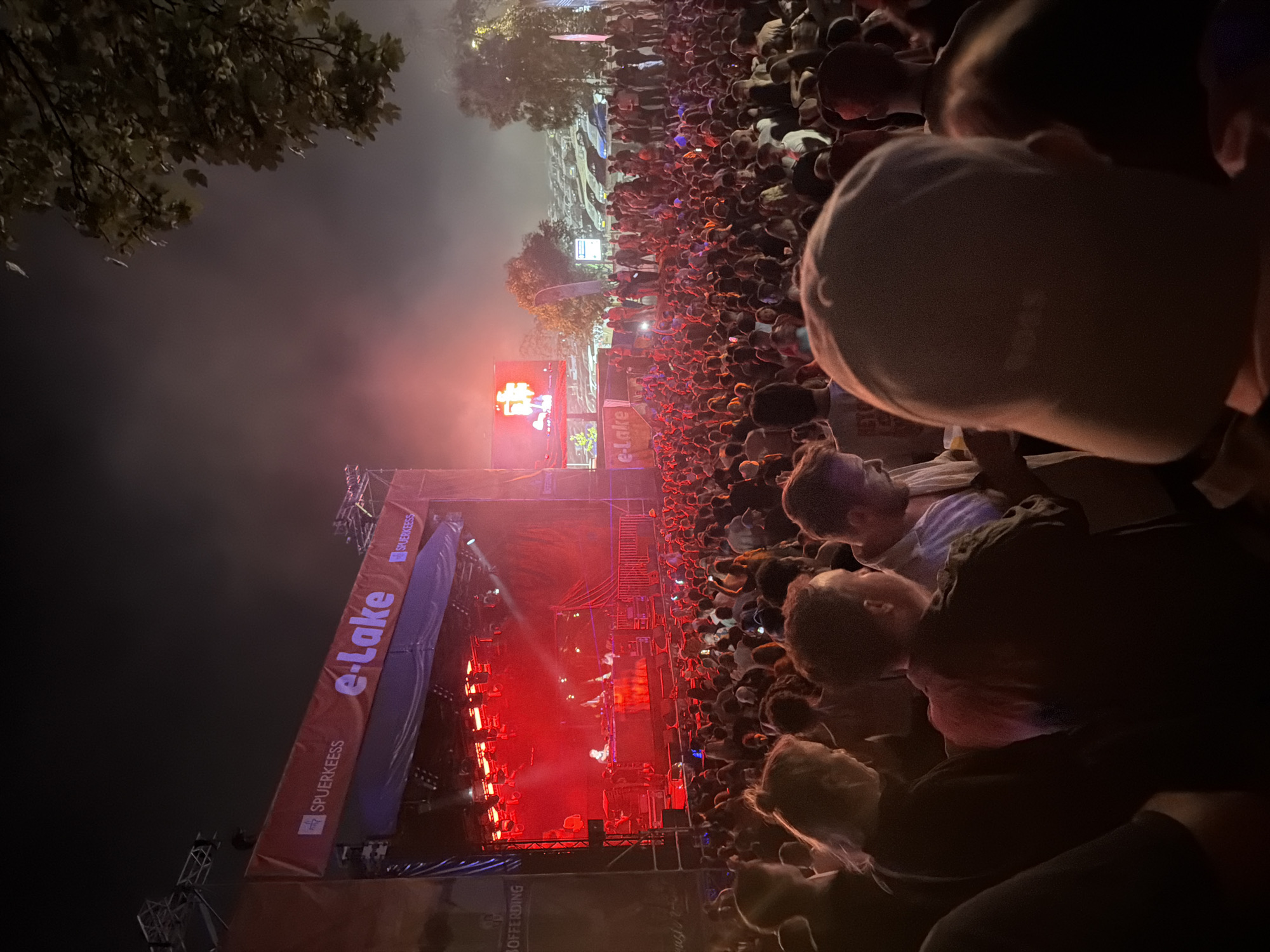 The image depicts a nighttime outdoor concert scene at the e-Lake festival in Echternach, Luxembourg. A large crowd of people is gathered in front of a stage illuminated by red lights. On the stage, several individuals appear to be performing or operating equipment. A large banner with the text "e-Lake" and "SPUERKEESS" is visible above the stage. In the background, a parking lot filled with cars and trees can be seen. The atmosphere is energetic, with the audience appearing engaged with the performance. The lighting is dramatic, with strong red hues dominating the scene.