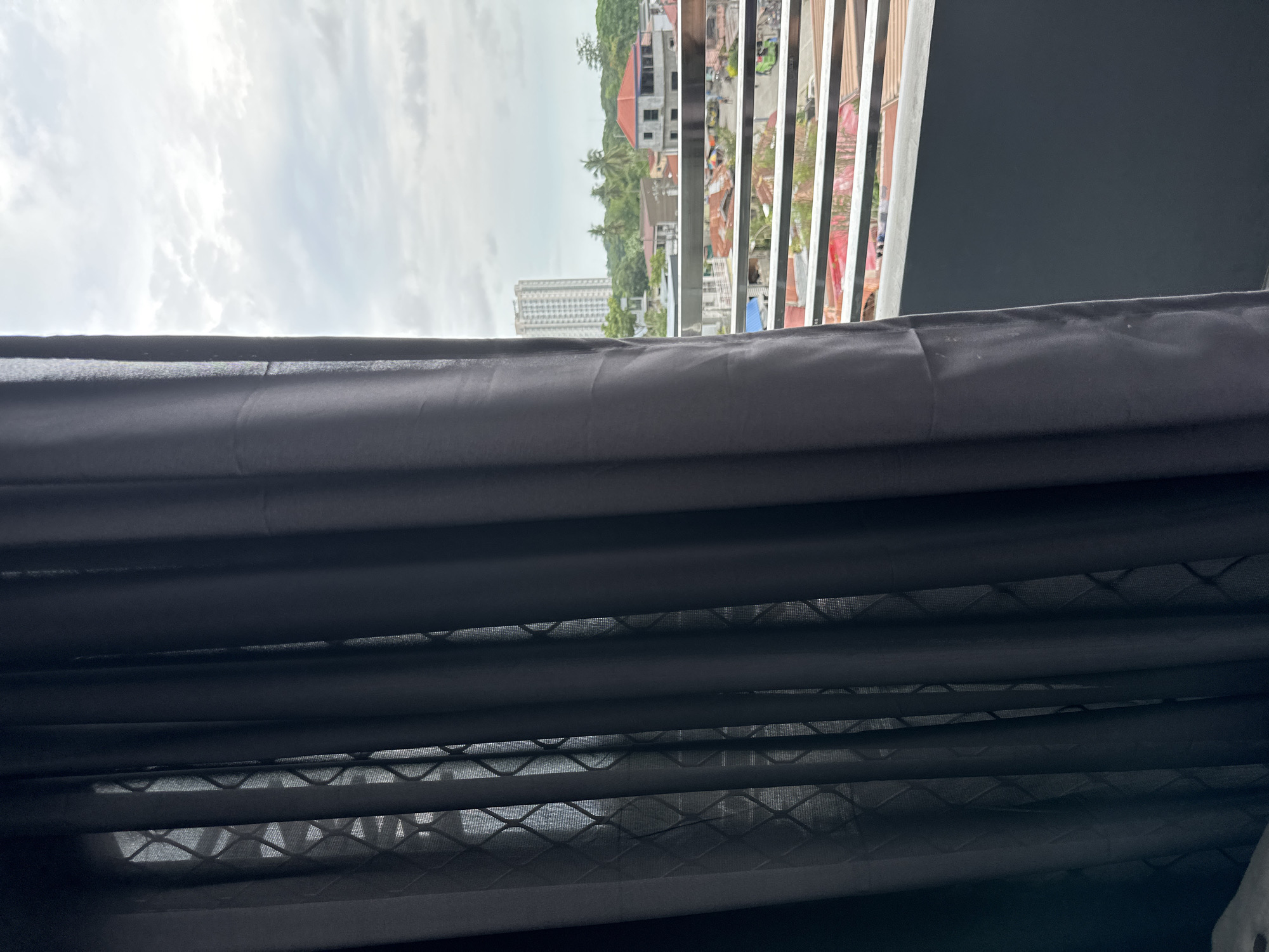 View from behind dark curtains in Davao City, Philippines. Outside the window, a cityscape is visible, including buildings, trees, and the sky. A tall building is located in the mid-ground. The curtain appears to be a combination of blackout and sheer.