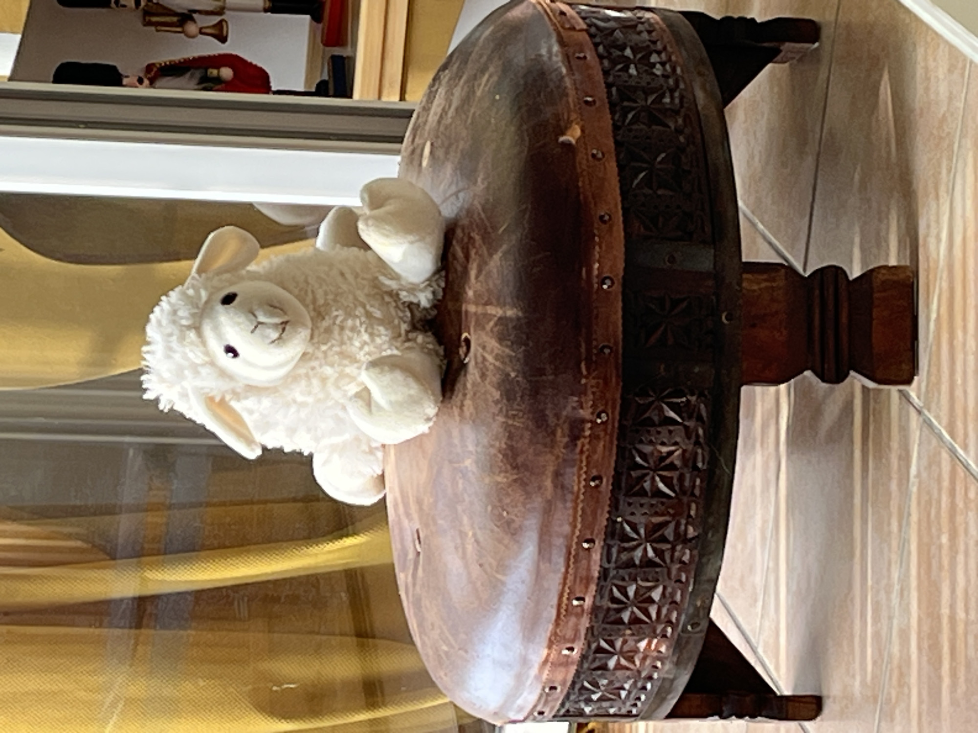 A stuffed sheep toy sits on a dark brown leather-topped ottoman with decorative carved wood sides. The ottoman has four carved wooden legs and is positioned on a tiled floor. In the background, a yellow curtain is visible, and through a window or glass partition, two nutcracker figures and a portion of a wooden shelf with items can be seen. The scene appears to be indoors, likely in a residential setting in Montélimar, France.