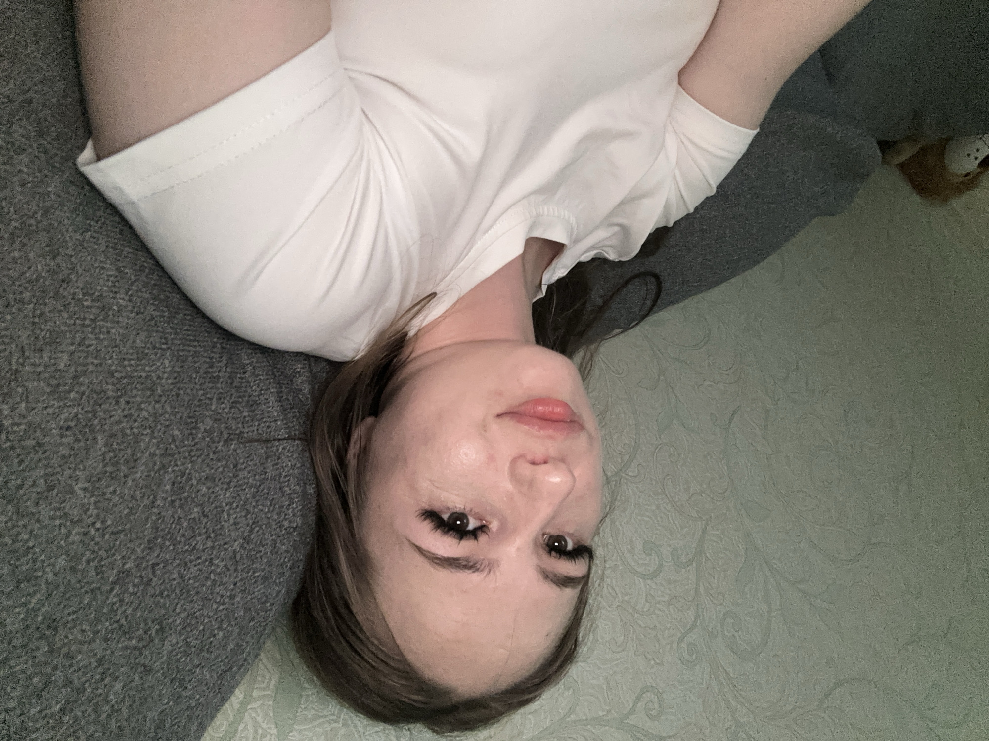 A young Caucasian woman is shown from the chest up, reclining on a grey couch. She has long brown hair, large false eyelashes, and is wearing a plain white t-shirt. She is looking slightly to her left, with her head resting on the couch. The background consists of textured, light green wallpaper on the left and the grey couch on the right. A stuffed lion toy is visible on the far left of the image, partially obscured by the couch. The lighting is dim, suggesting an indoor setting.