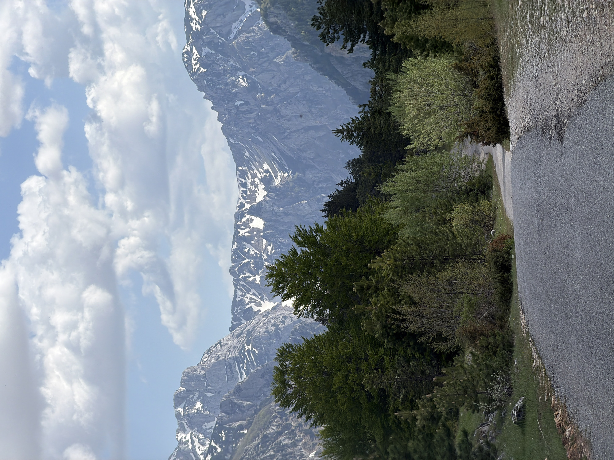 The image shows a mountain landscape in Mala Crna Gora, Montenegro. The scene includes a paved road running along the right side of the frame. To the left of the road, there is lush green vegetation, including trees and shrubs. In the background, a large, snow-capped mountain range dominates the view, partially obscured by clouds. The sky is blue with scattered white clouds.