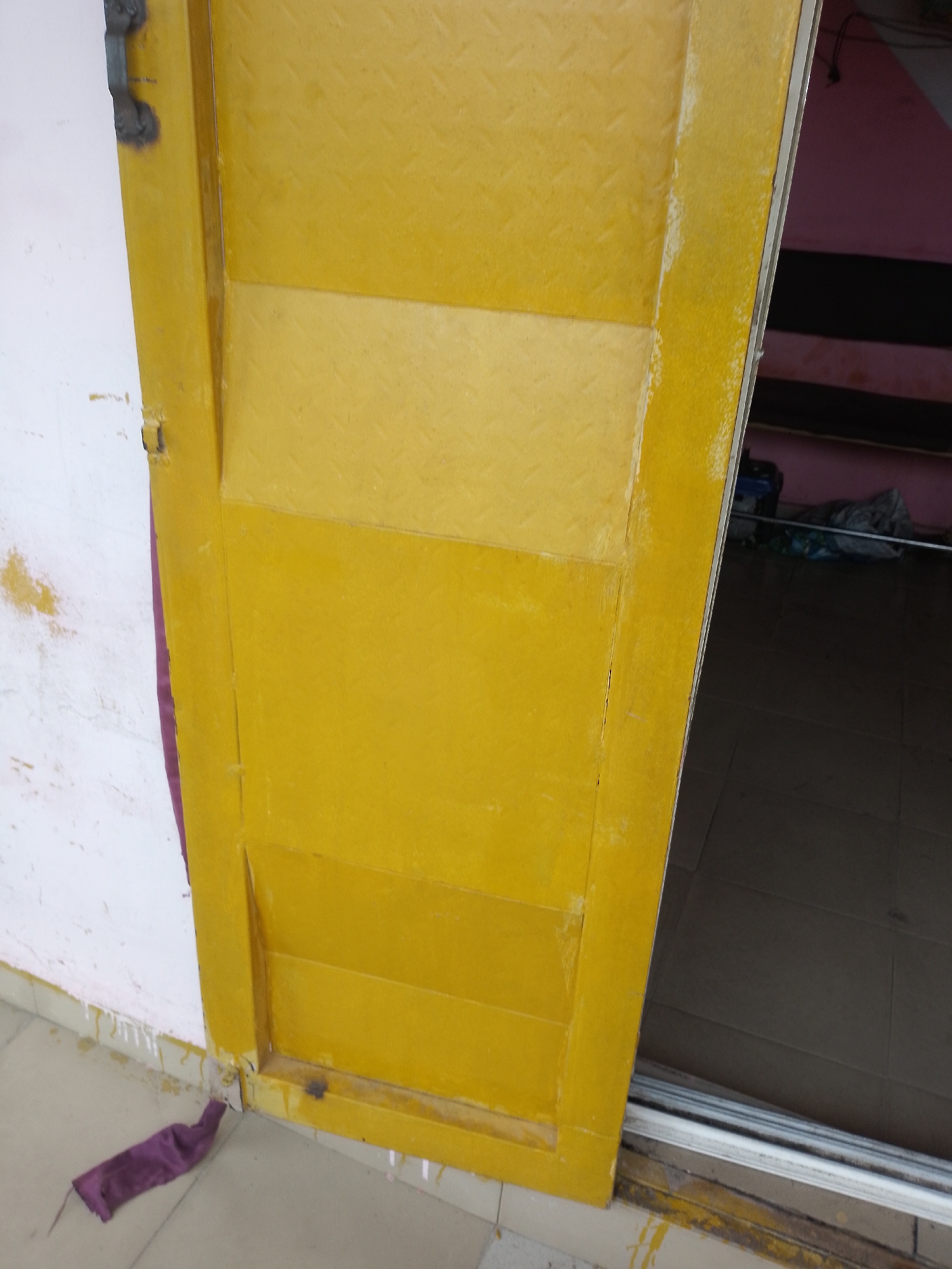 An open yellow metal door is visible from a side angle, revealing an interior space. The door has a textured surface with a diamond pattern. The wall to the left of the door is white and appears worn with some yellow discoloration. A piece of dark purple fabric is draped over the bottom edge of the door and rests on the light-colored floor. The interior space visible through the doorway has a tiled floor and shelves with various objects, including bags and what appears to be a blue container. The scene suggests a utilitarian or storage area.