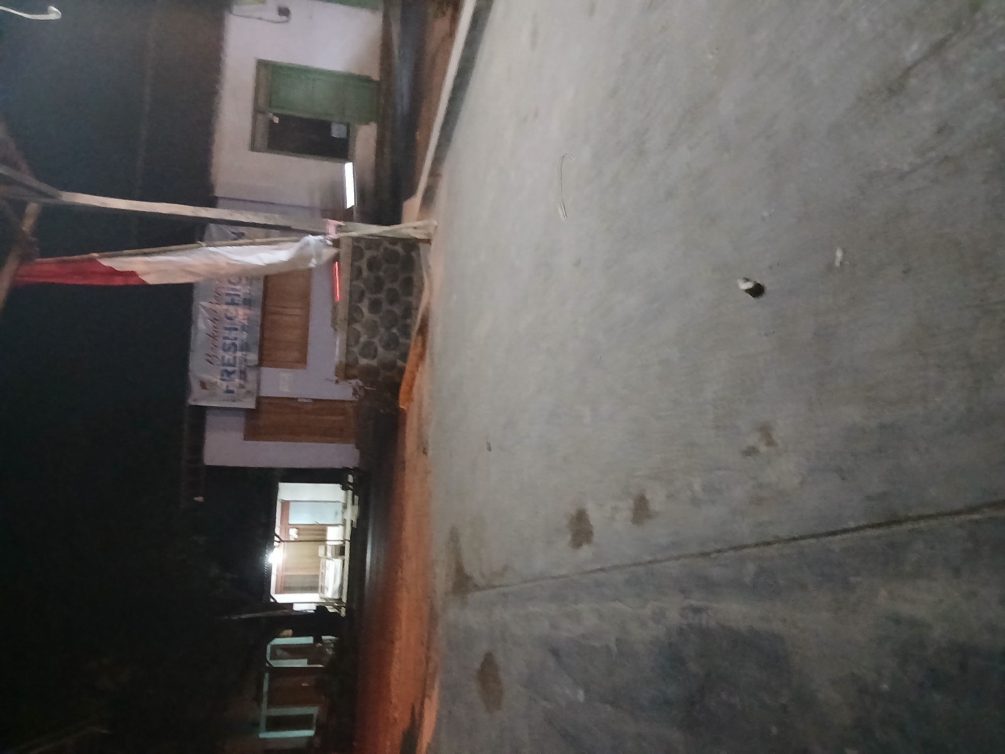 The image shows a street scene at night in Tenggarang, Indonesia. In the background, there are buildings with lights on, suggesting they are businesses or residences. A red and white Indonesian flag is visible, hanging from a pole. The foreground shows a paved street with some visible stains or puddles. The overall impression is of a dimly lit, possibly rural or suburban area at night.
