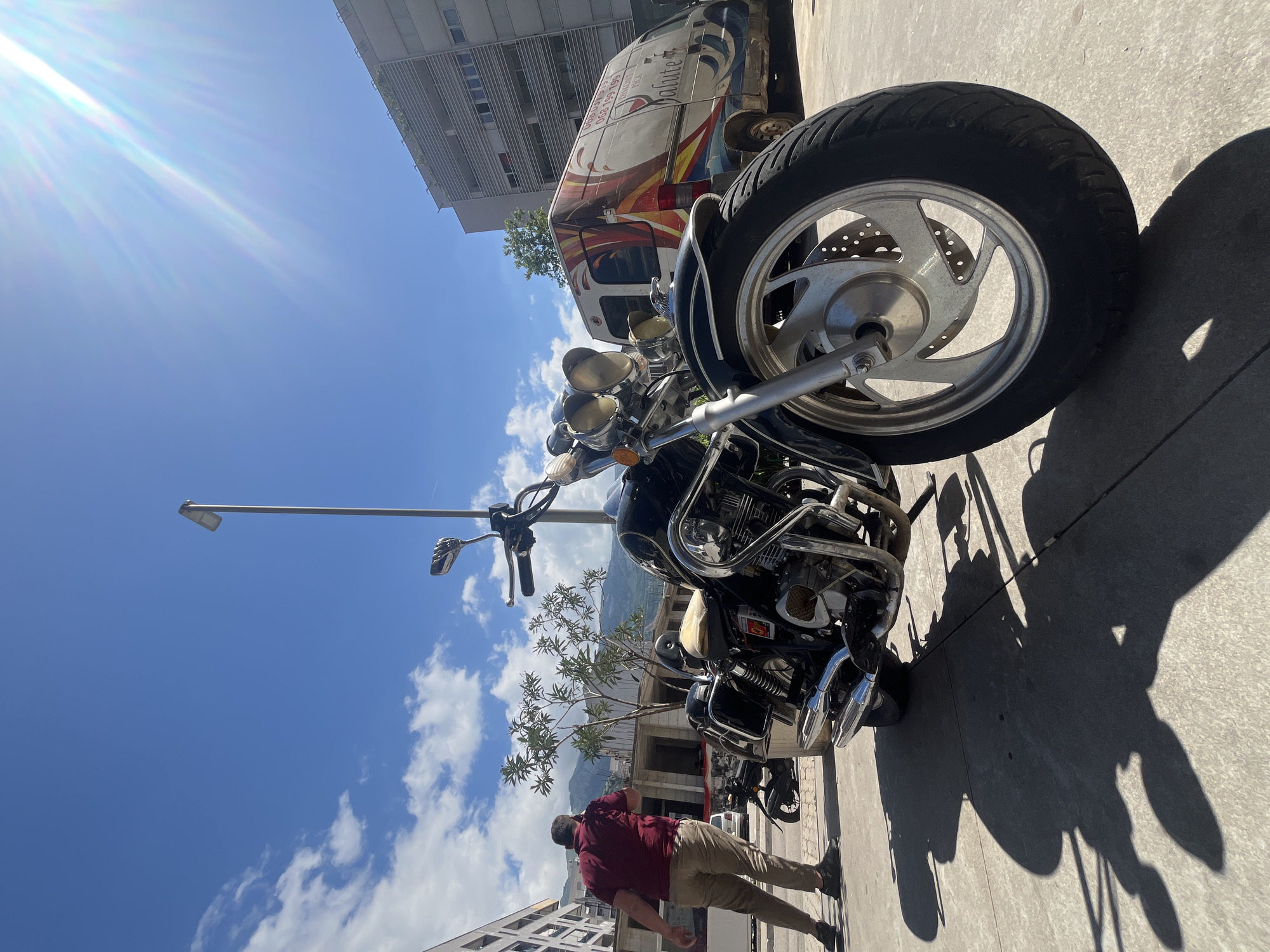 The image, if viewed with correct orientation, depicts a bright street scene in Budva, Montenegro, under a clear blue sky. In the foreground on the right, a large, black cruiser-style motorcycle is parked, showcasing its chrome engine components, multiple headlights, and a robust front wheel with a disc brake. Behind the motorcycle, a white delivery van with vibrant, swirling graphics in red, orange, and blue is visible. Text on the van includes "UNE GASTRONOMIE RICHE ET VARIEE," "Saluté," and a phone number "063 591 091."
In the lower-left section of the frame, a man wearing a maroon collared shirt and khaki pants walks away from the viewer. Tall, modern buildings line the street on the left, and a streetlamp stands further back. In the midground, green trees are visible, with distant mountains discernible beneath the horizon. Strong sunlight emanates from the upper left corner of the sky, creating a significant lens flare and casting long, distinct shadows of the motorcycle and the man onto the light-colored concrete pavement, suggesting a mid-morning or early afternoon setting on a sunny day with scattered white clouds.