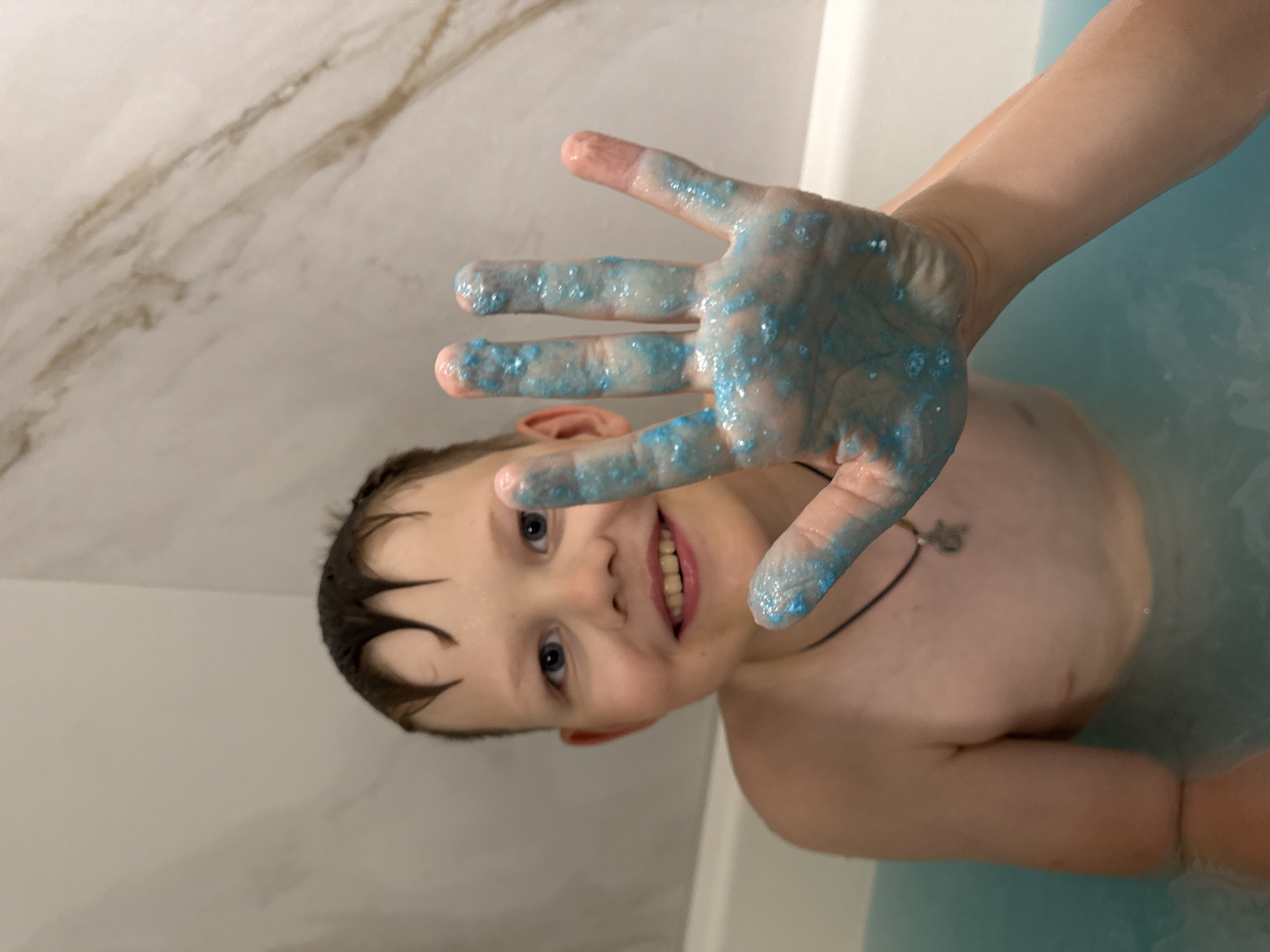 A young boy with dark, wet hair and blue eyes is in a bathtub filled with blue water. He is smiling and looking at the camera. His right hand is held up in front of him, with blue glitter or bath bomb residue covering his palm and fingers. The bathtub has white walls with a marbled pattern and a white rim. The boy is wearing a dark necklace with a silver charm. The scene appears to be a private bathroom. The location in the city of Ферма, Russia is not discernible from the image.
