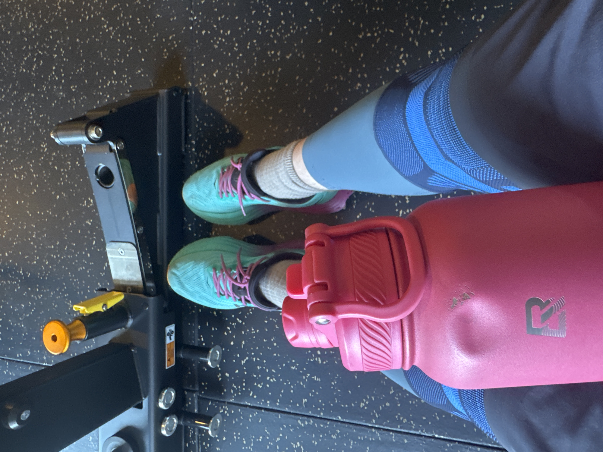 This overhead shot captures the lower body of a person at a gym located in Lâm Viên - Đà Lạt, Vietnam. The individual is wearing light blue athletic leggings with a prominent darker blue patterned design, light grey socks, and vibrant teal-colored athletic shoes with purple laces. Their feet are positioned on a dark, speckled rubber gym floor, which is characteristic of fitness facilities.
Adjacent to the person's feet is a robust piece of black gym equipment, likely part of a weight-training machine such as a squat rack. This equipment features yellow and orange adjustable components, along with various metallic bolts and a small, unreadable warning sticker. Resting next to the person is a large, bright pink water bottle. The bottle shows signs of use, including a visible dent and scuff marks, and features a silver, stylized 'R' logo prominently on its side.
The scene suggests a moment of rest or preparation during a workout in an indoor fitness environment. The limited view of the person precludes any interpretation of emotions, and the time of day is not discernible.