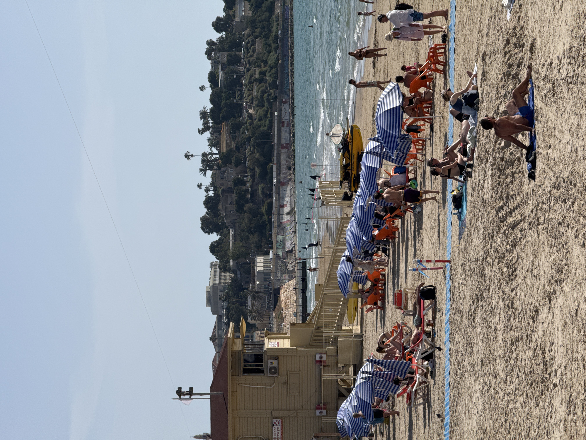 The image shows a beach scene in Tel Aviv, Israel. The foreground is dominated by a sandy beach, populated with beachgoers sitting in chairs under blue and white striped umbrellas. There is a blue rope lying along the border. The background includes the Mediterranean Sea and a hillside dotted with buildings and vegetation. A lifeguard station is visible near the foreground. People are seen swimming and walking along the shoreline.