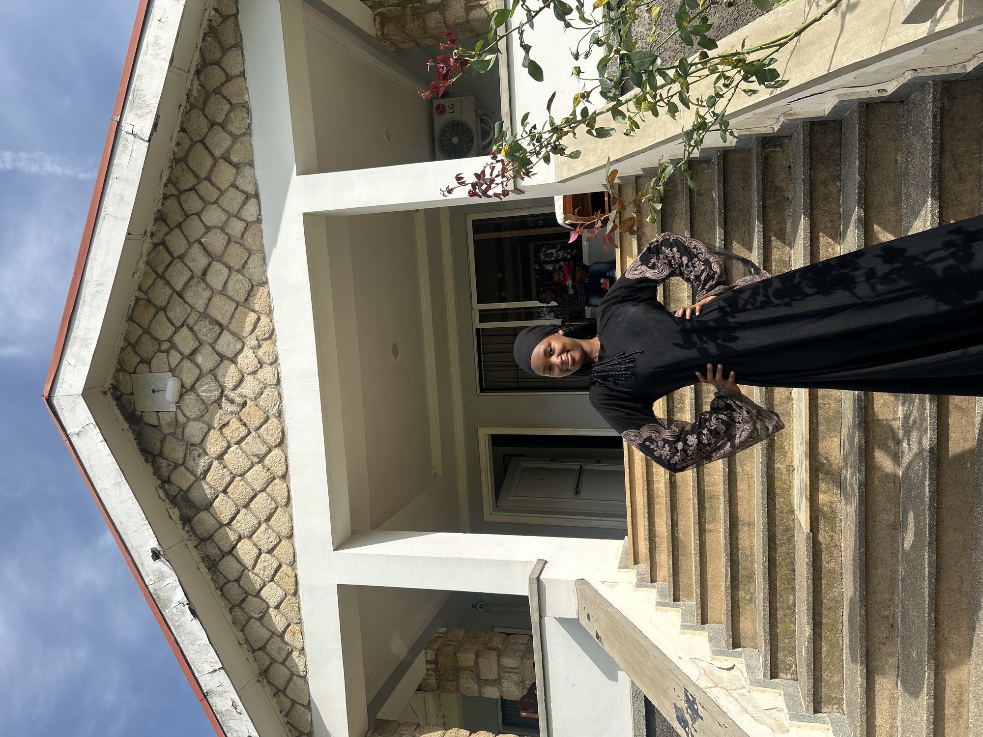 A young woman wearing a black hijab and a black abaya with embroidered sleeves stands on a concrete staircase outside a building. The building's facade features a triangular section with a stone-tiled pattern. To the left, the sky is blue with wispy clouds. A rose bush with red and green leaves hangs over the scene from above. An air conditioning unit is visible on the wall of the building. The woman is smiling and looking at the camera, with her hands on her hips. The lighting suggests it is daytime. The overall setting appears to be residential.